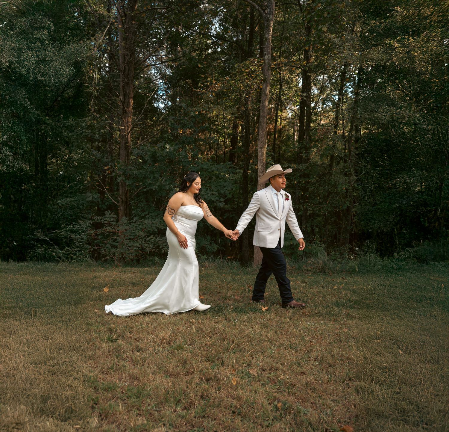 Bride and groom in white cowboy hat hold hands walking across green lawn with forest backdrop.