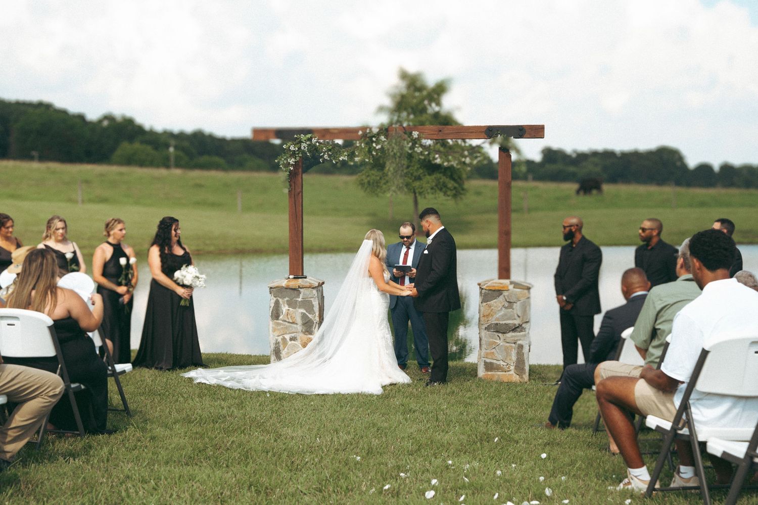 Outdoor wedding ceremony by a pond, bride and groom at wooden arch altar with wedding party and seated guests on green lawn.