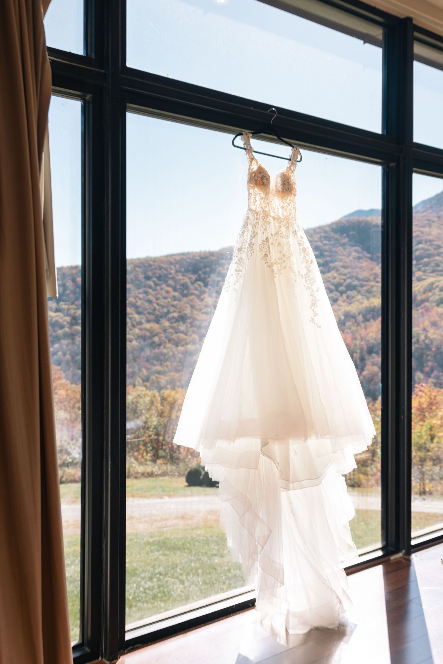 Elegant lace wedding gown hanging on a hanger in front of large windows with autumn mountain views.