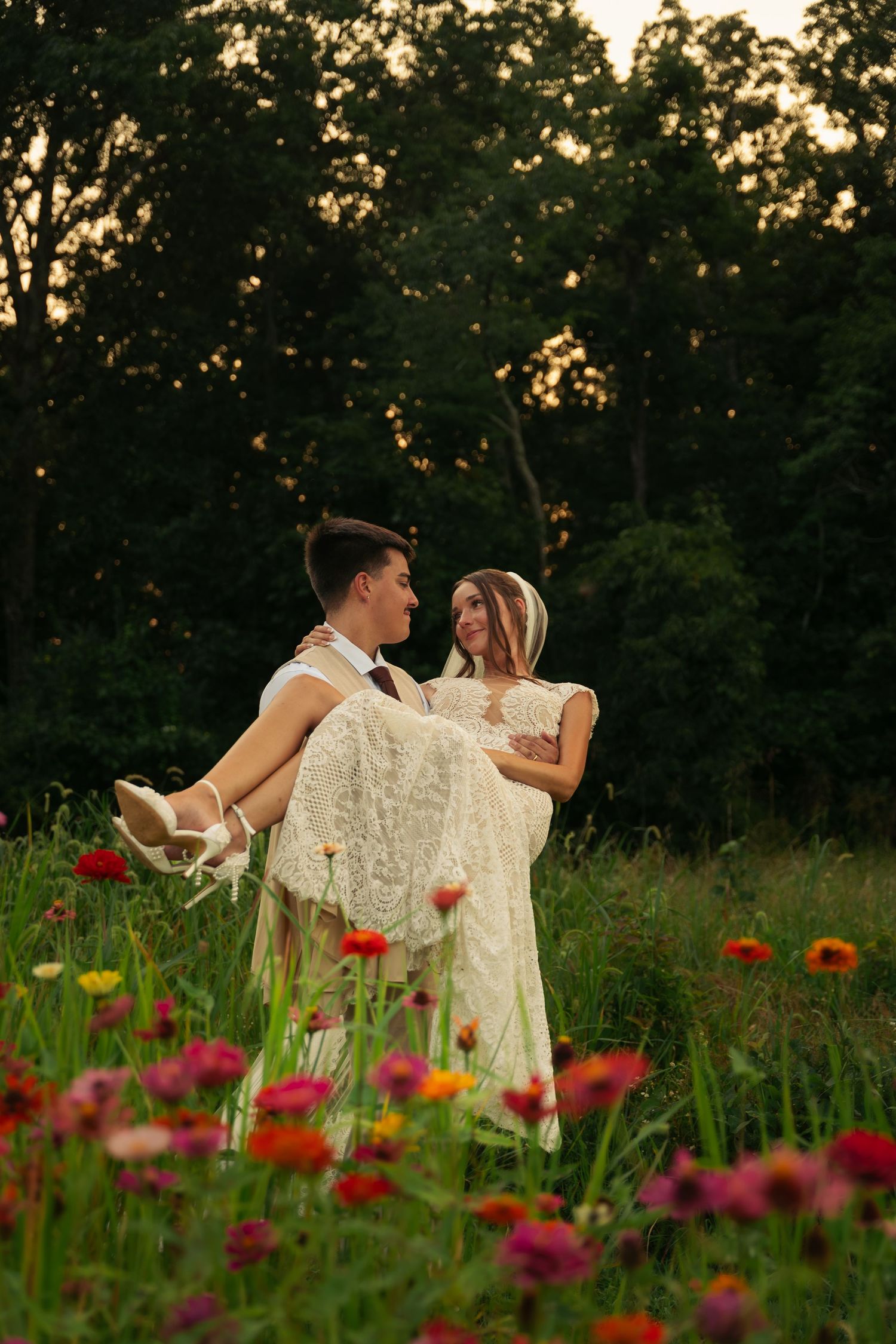 Groom carrying bride in white lace dress through a colorful flower field at sunset with trees in background.