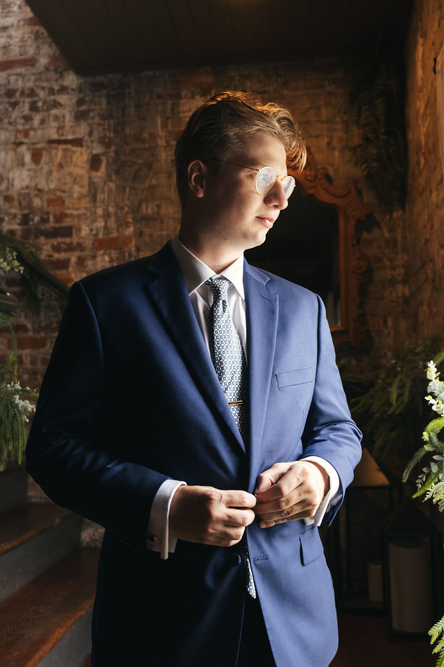 Young redhead groom in blue suit buttoning jacket indoors with brick wall and greenery backdrop.