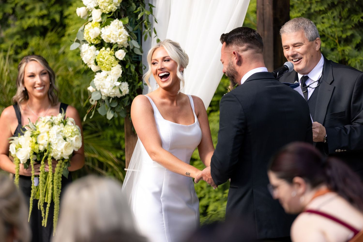 Bride laughing joyfully during outdoor wedding ceremony, holding hands with groom, bridesmaid holding white bouquet nearby.