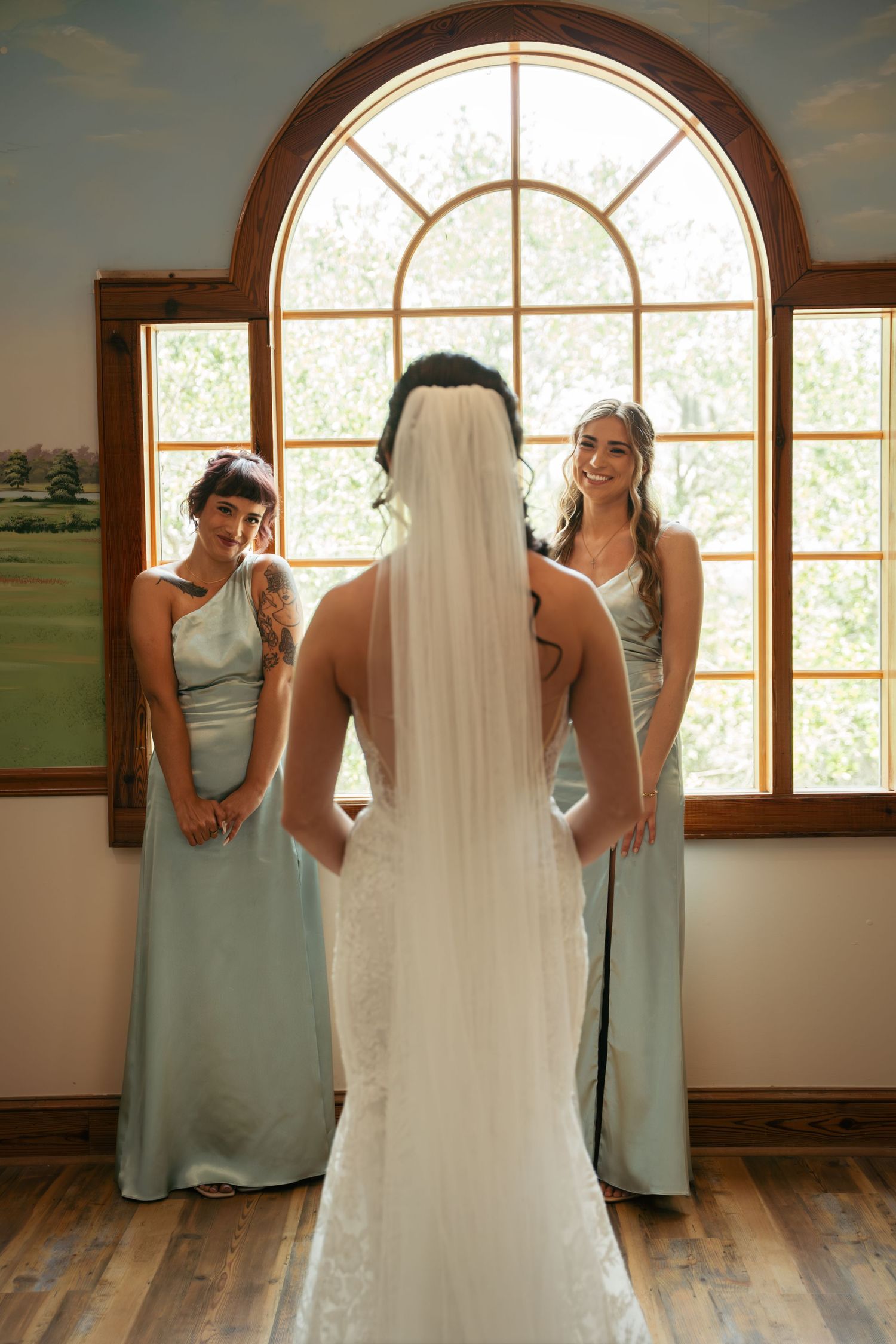 Bride in white gown and veil stands between two smiling bridesmaids in sage green dresses before an arched window.