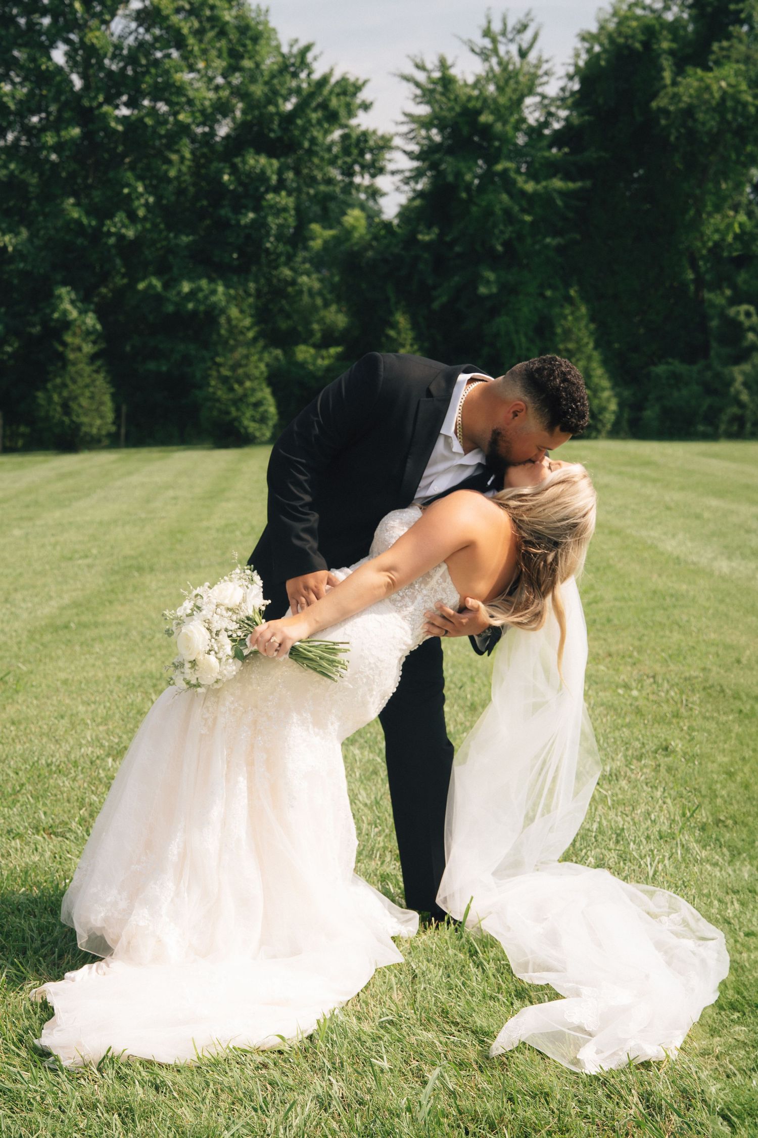Bride and groom share a romantic dip kiss on lush green lawn, bride in white lace gown holding bouquet.