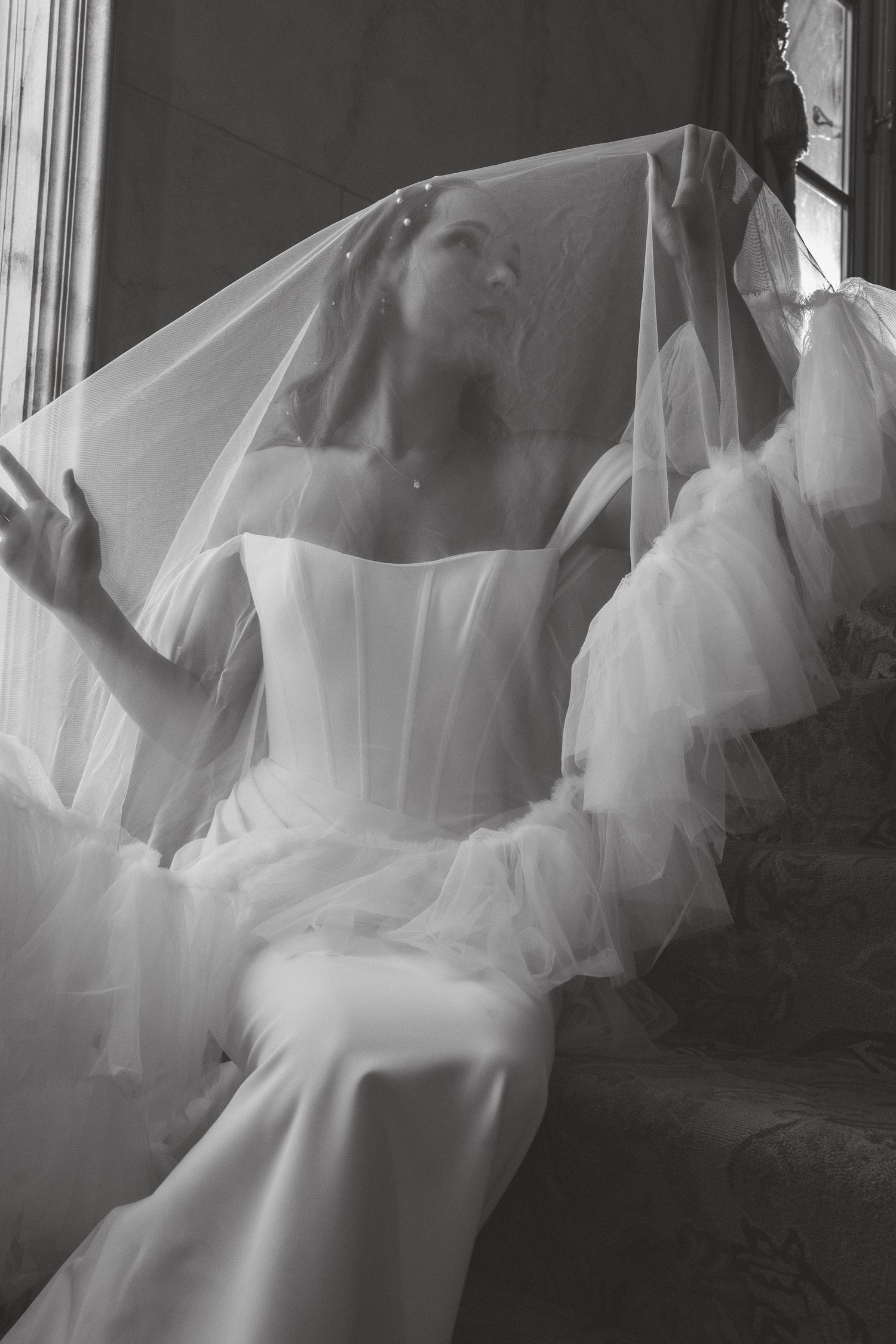 Black and white photo of a bride in a white corset gown and veil, seated gracefully on stairs.