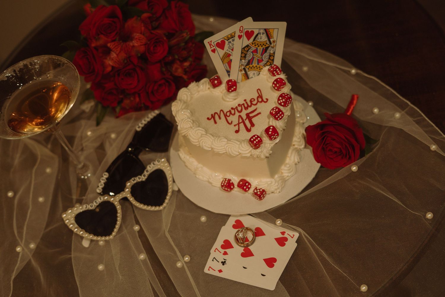 Heart-shaped wedding cake reading Married AF with playing cards, red roses, heart sunglasses, and dice on a glass table.