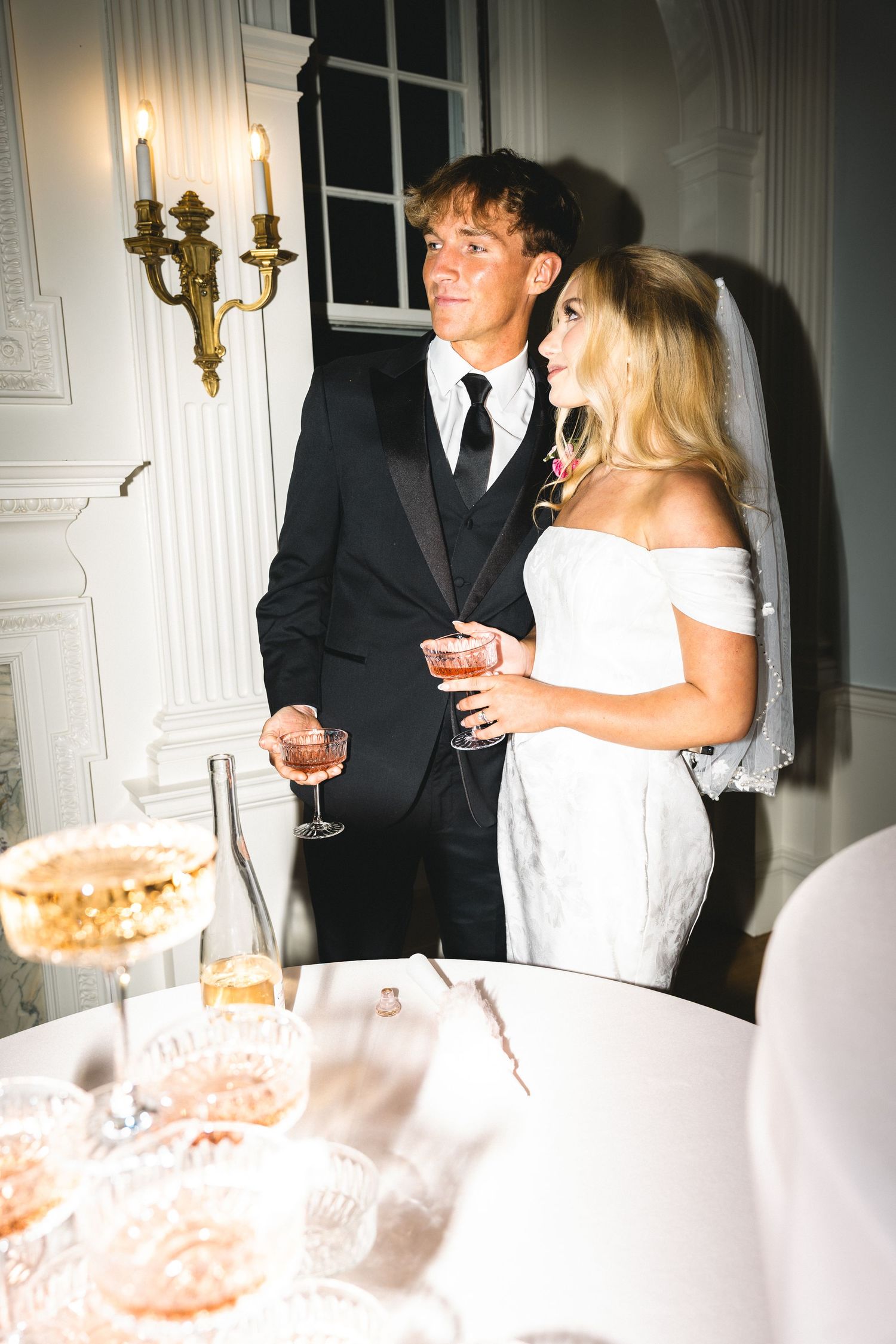 Elegant bride and groom holding champagne glasses at a wedding reception in a white ornate venue.