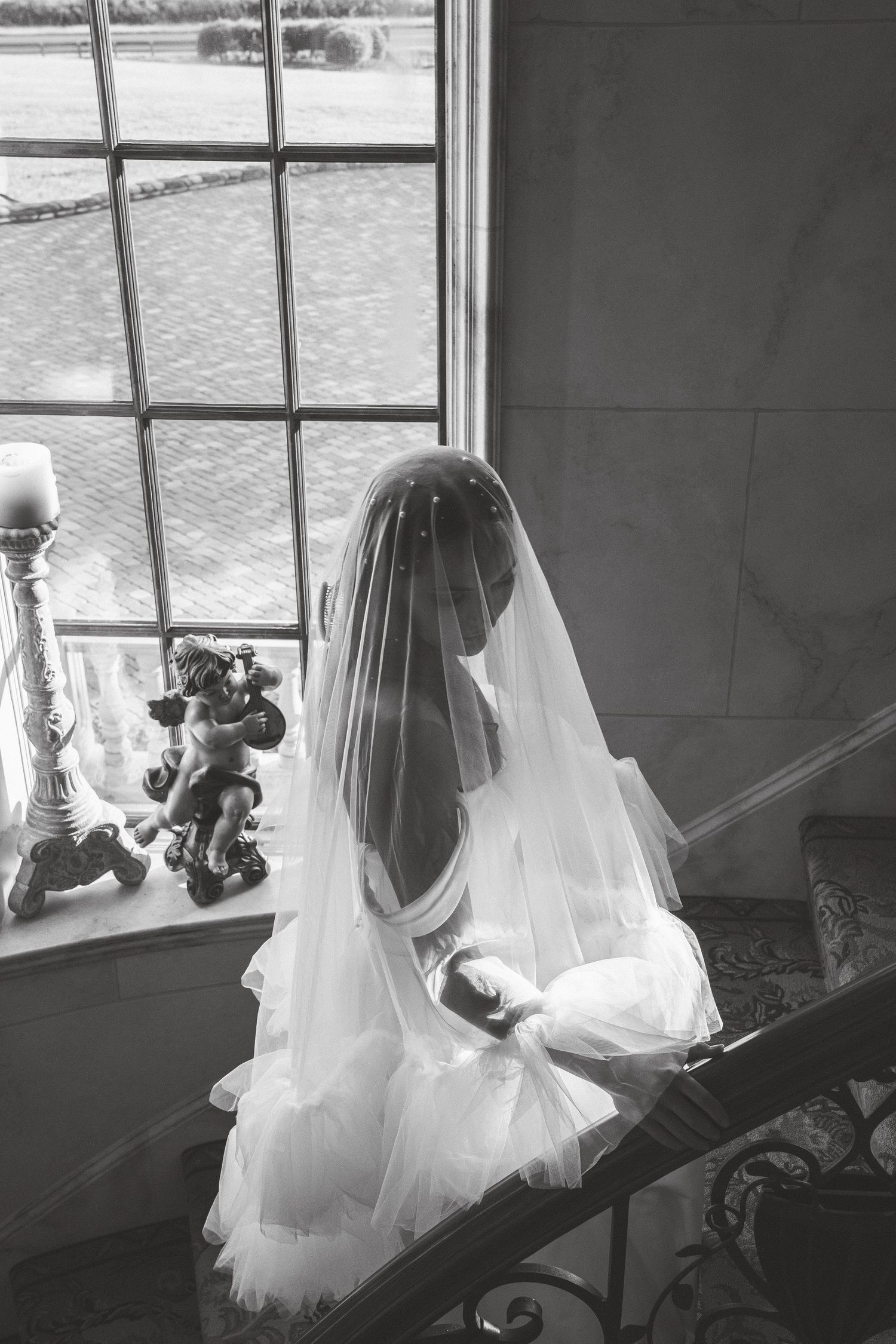 Elegant bride in veil sits on staircase by large window in black and white wedding portrait.