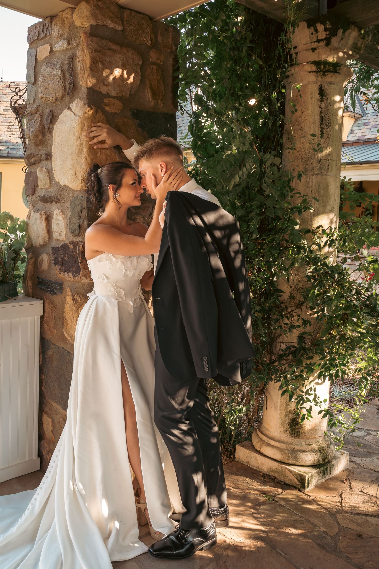Bride in strapless white gown and groom share romantic moment by stone columns draped in lush greenery.