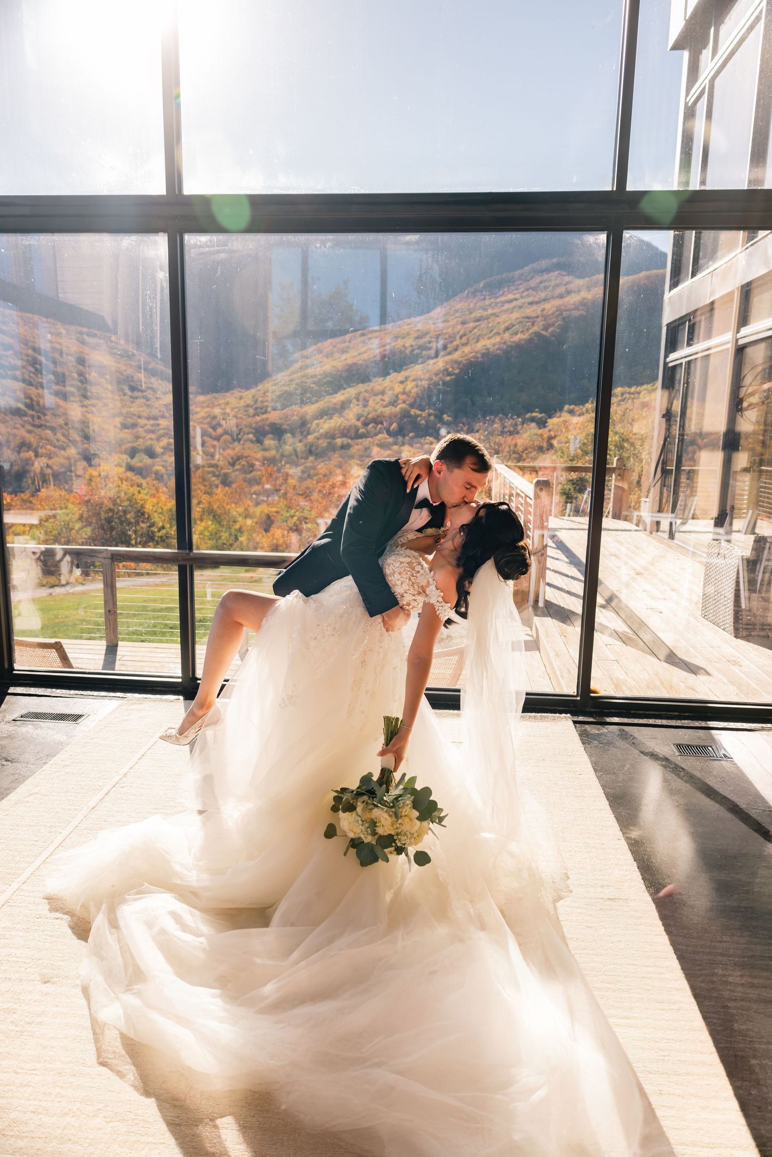 Bride and groom share romantic dip kiss indoors with stunning autumn mountain view through floor-to-ceiling windows.