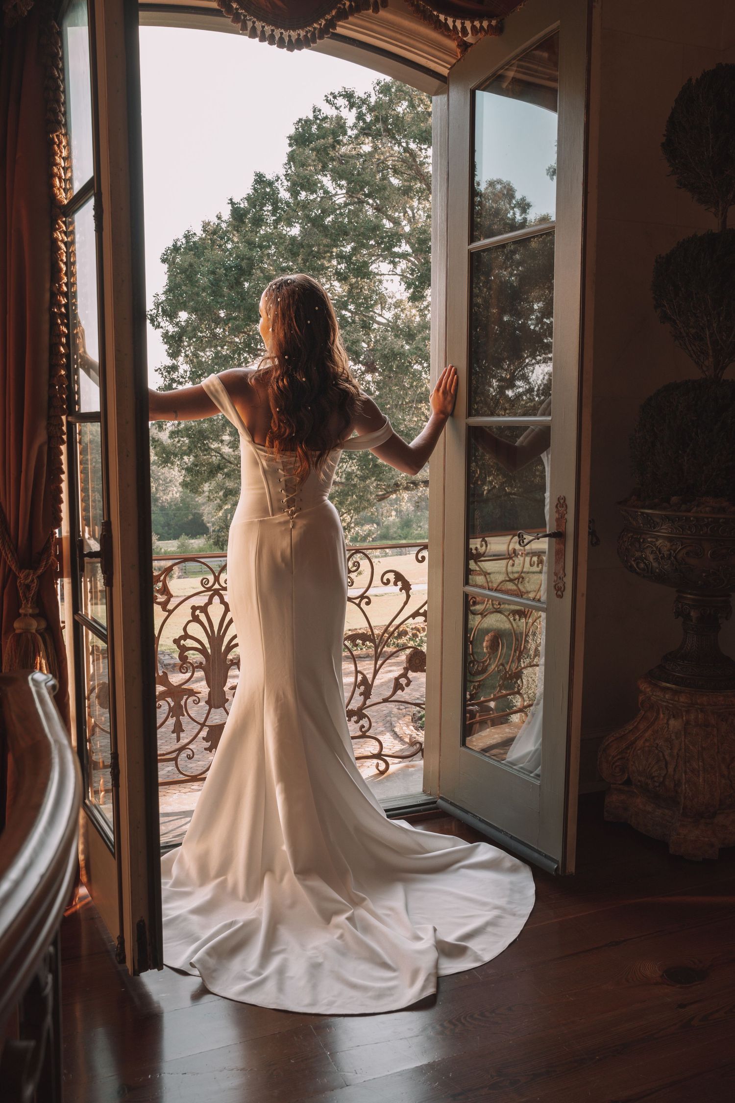 Bride in elegant white mermaid gown stands at ornate French doors overlooking lush garden balcony.