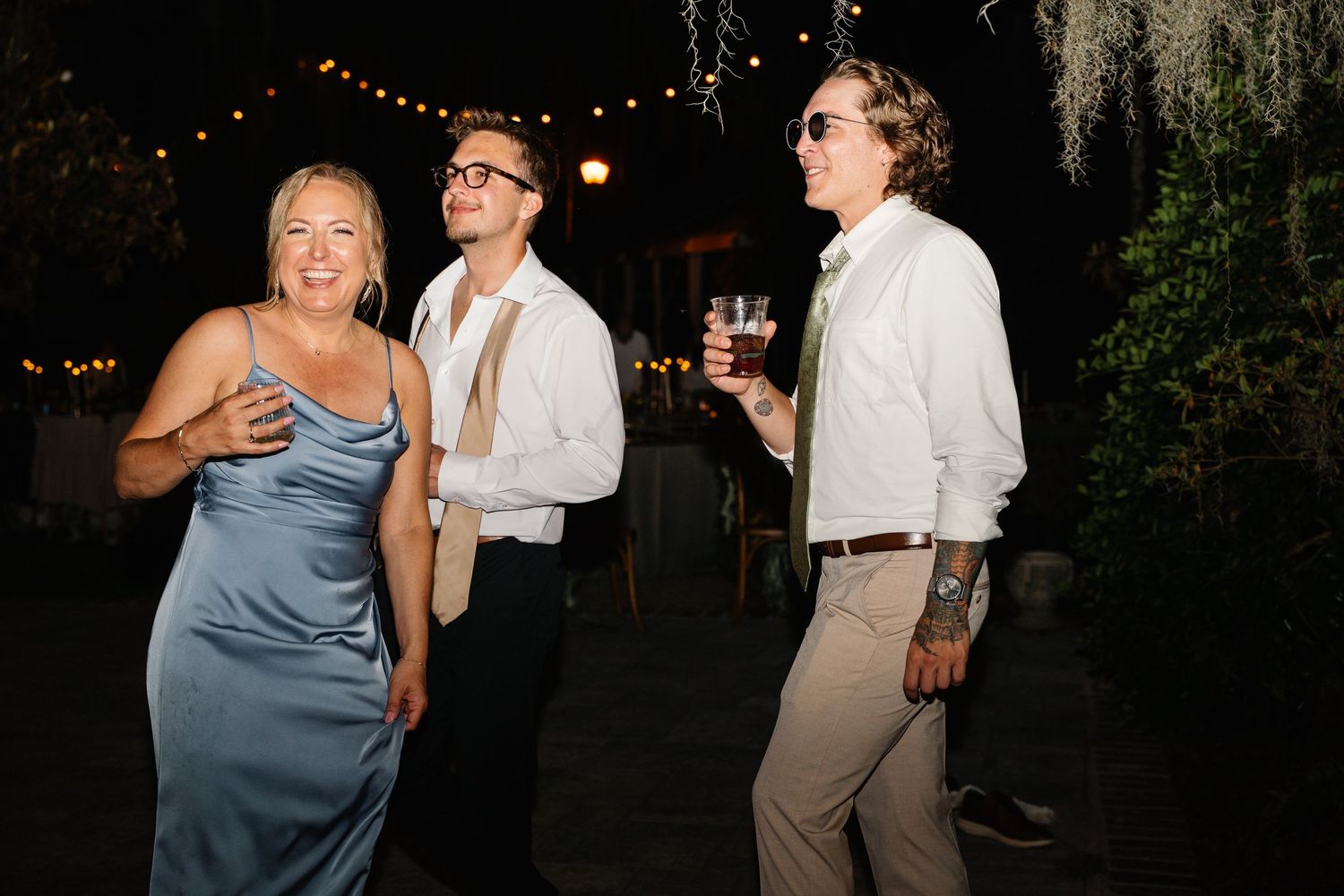 Three guests socializing at an outdoor evening wedding reception, wearing elegant formal attire.