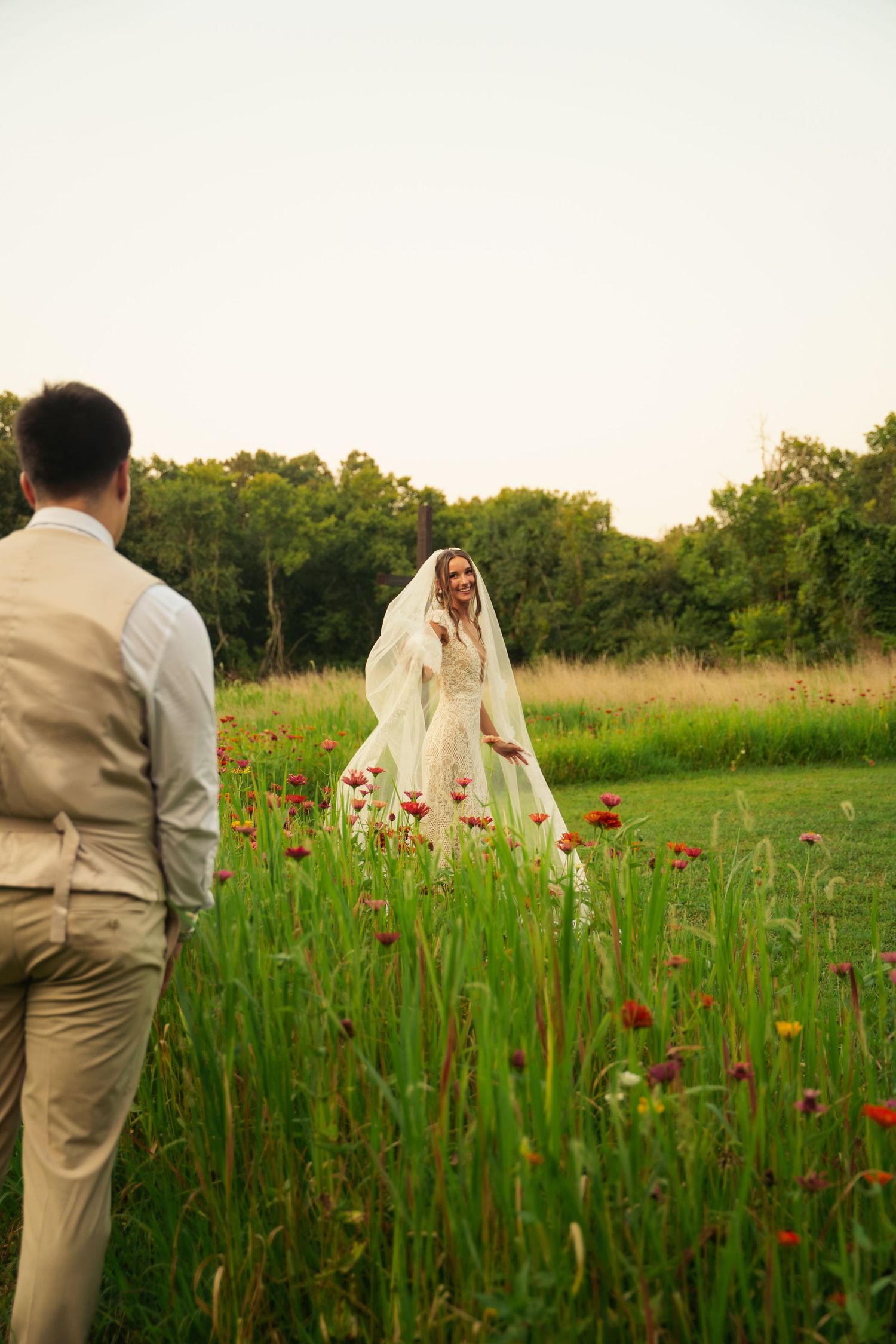 Bride in white gown and veil stands among red wildflowers, smiling at groom in tan suit in a lush green field.