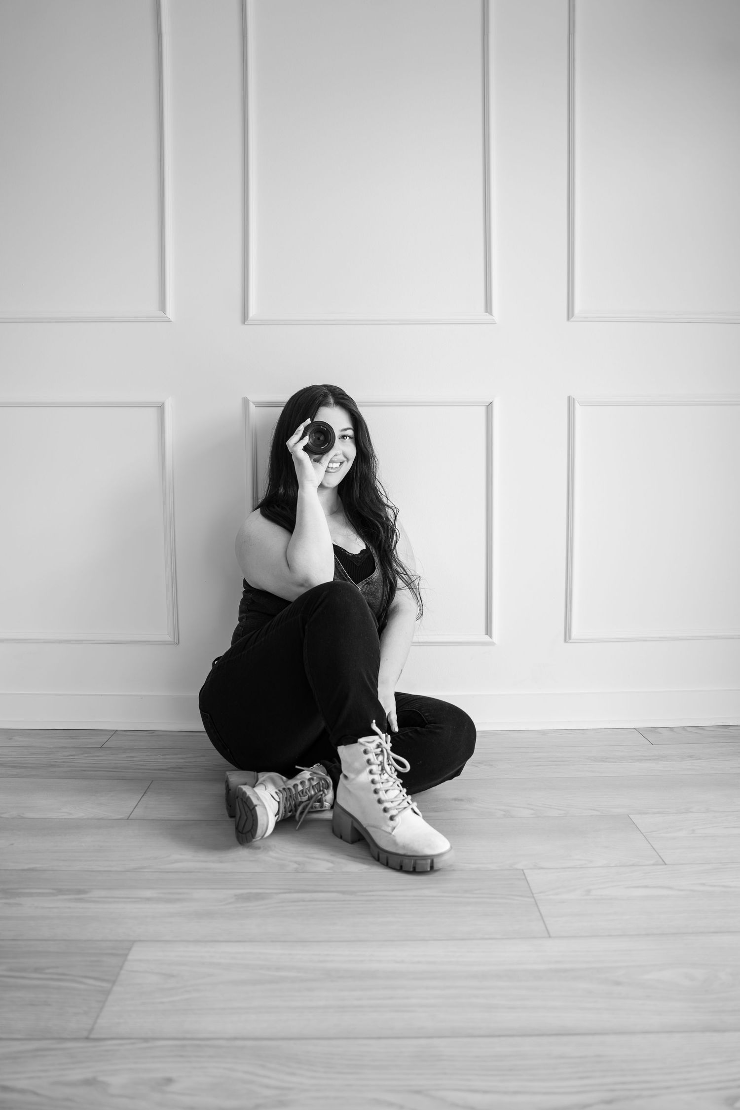 Black and white photo of a young woman sitting on a floor, holding a camera lens up to her eye, wearing boots.