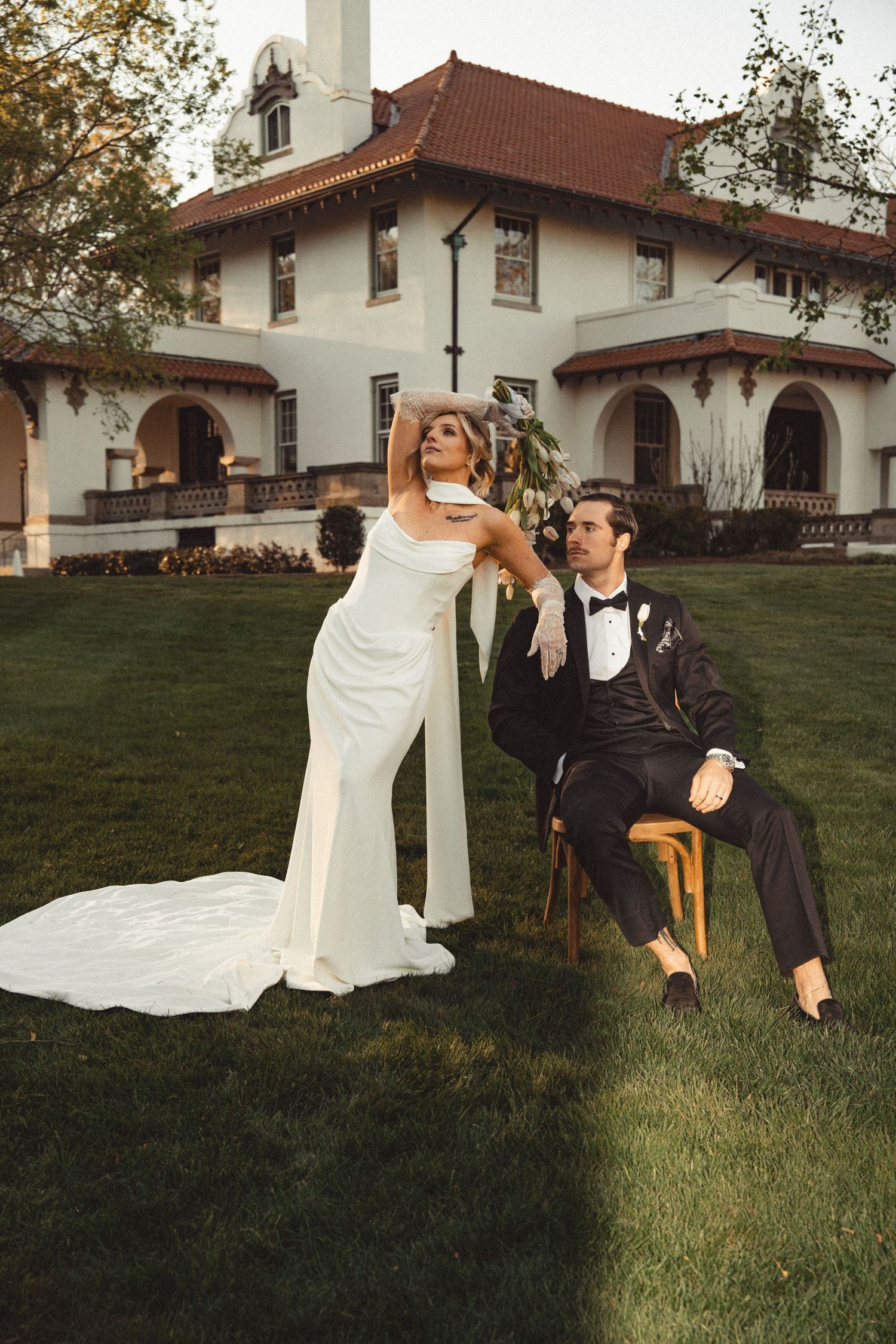 Bride in white gown and groom in black tuxedo pose elegantly on lawn before a grand white Spanish-style mansion.