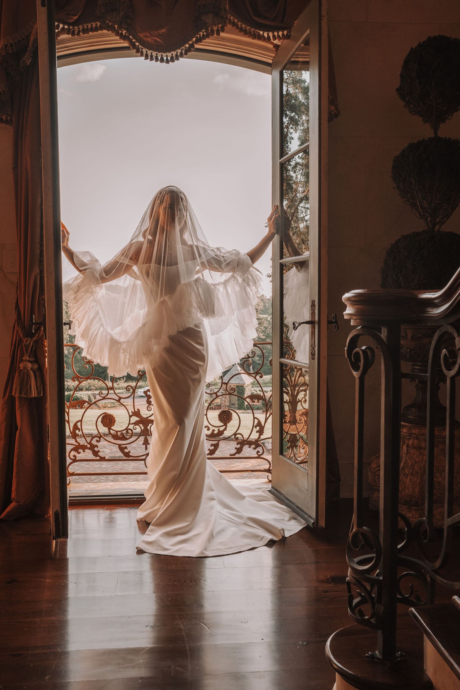 Bride in white gown and veil stands in ornate doorway opening to balcony with scenic view.