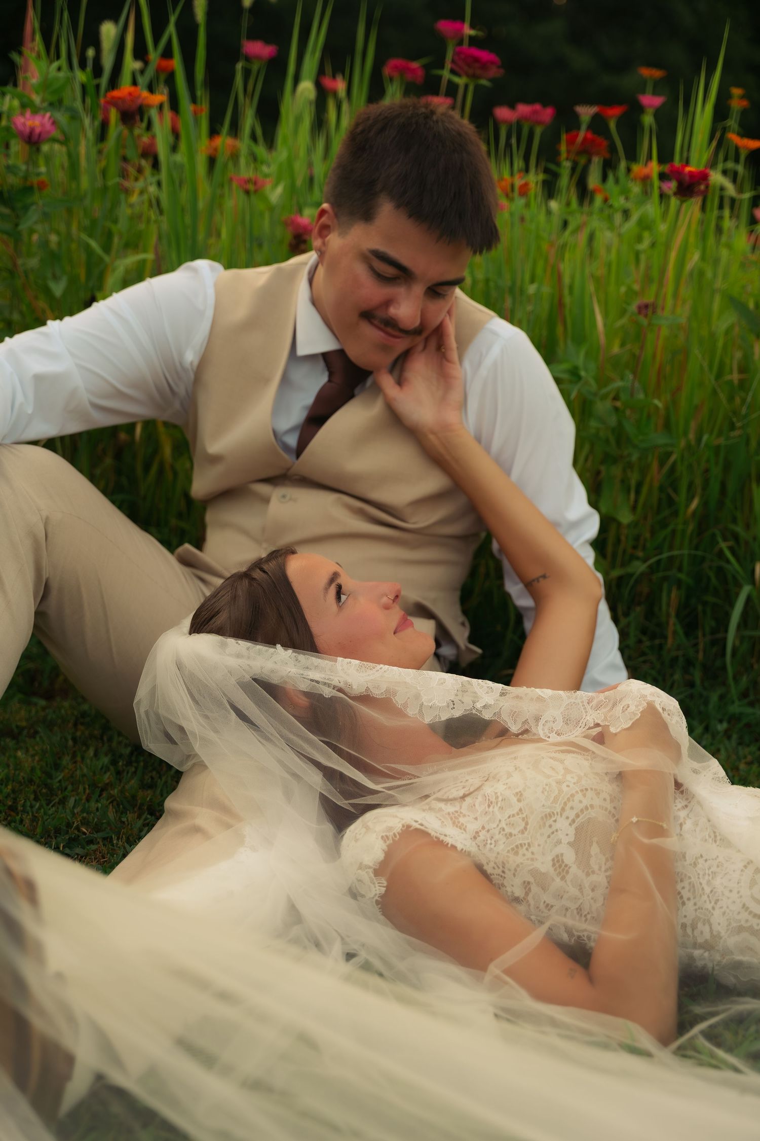 Bride and groom sitting in garden, bride lying in groom's lap wearing lace dress and veil, surrounded by colorful flowers.