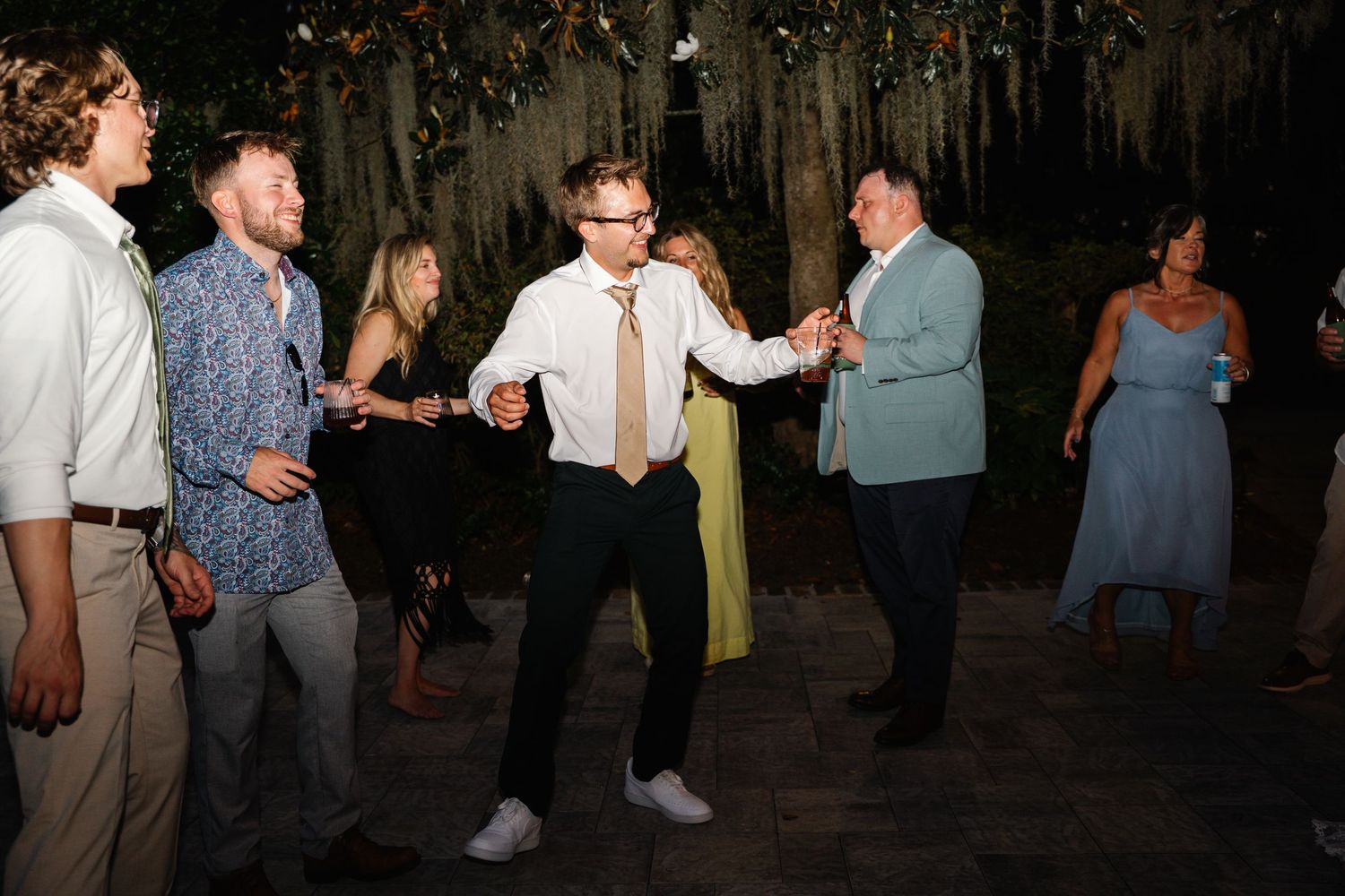 Guests dancing energetically at an outdoor wedding reception at night, surrounded by lush greenery and string lights.