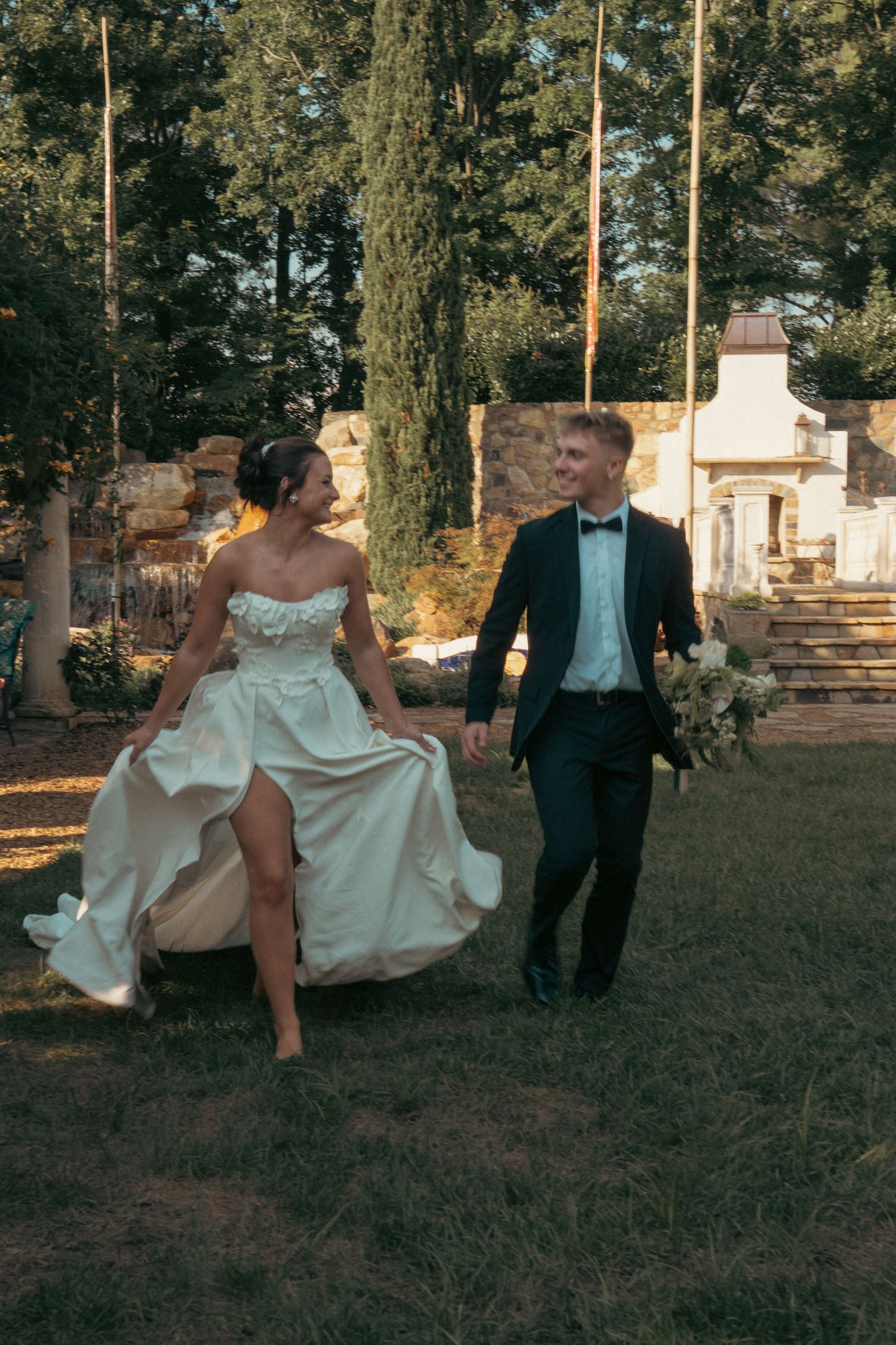 Bride and groom walking hand in hand outdoors, laughing joyfully, groom holding bouquet, lush garden wedding venue backdrop.