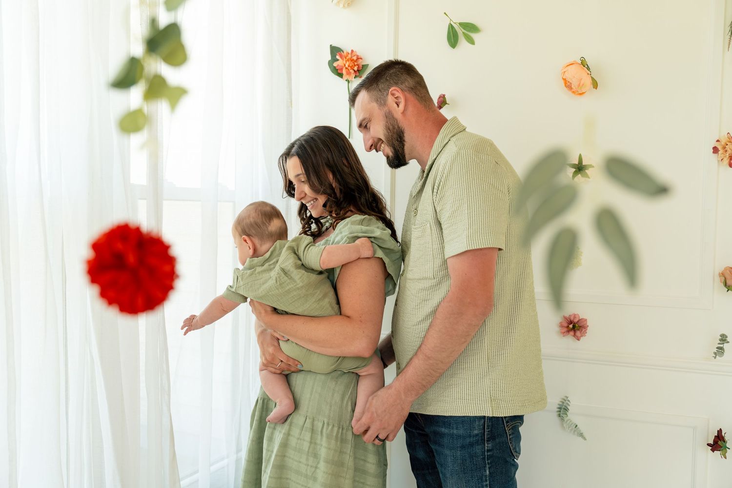 A young family with a newborn baby poses in a bright studio with floral decorations and green outfits.