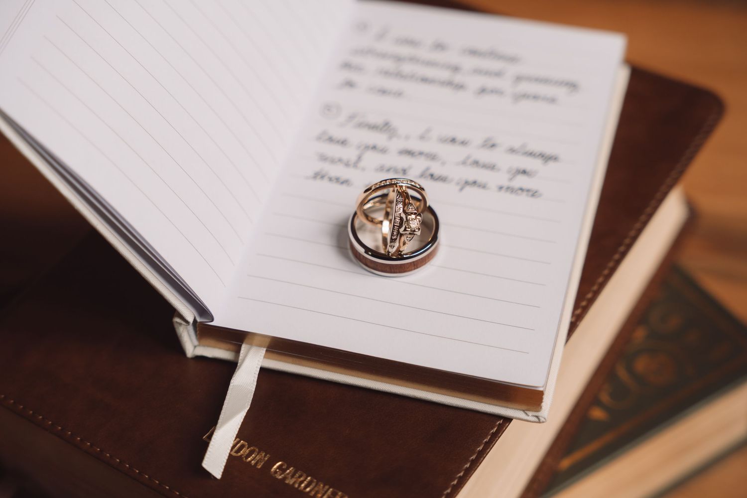 Wedding rings resting on an open journal with handwritten vows, placed on a leather-bound book.