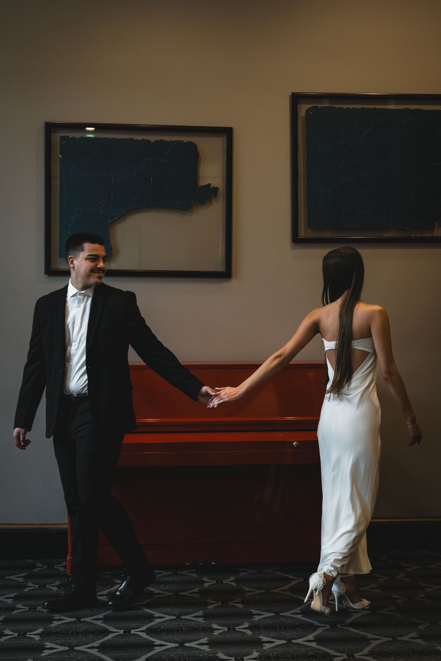 Elegant couple holding hands near a vintage upright piano, man in black tuxedo and woman in white backless gown.