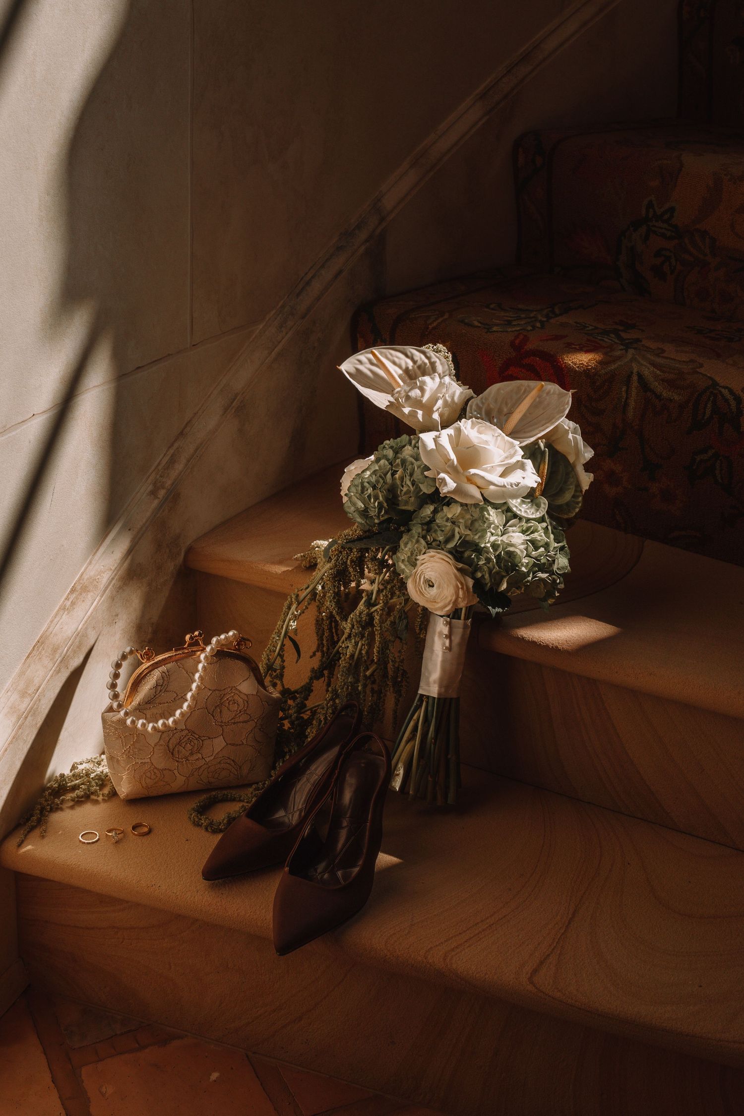 Wedding bouquet with white roses, bridal heels, pearl clutch, and rings arranged on warm-toned stairs.