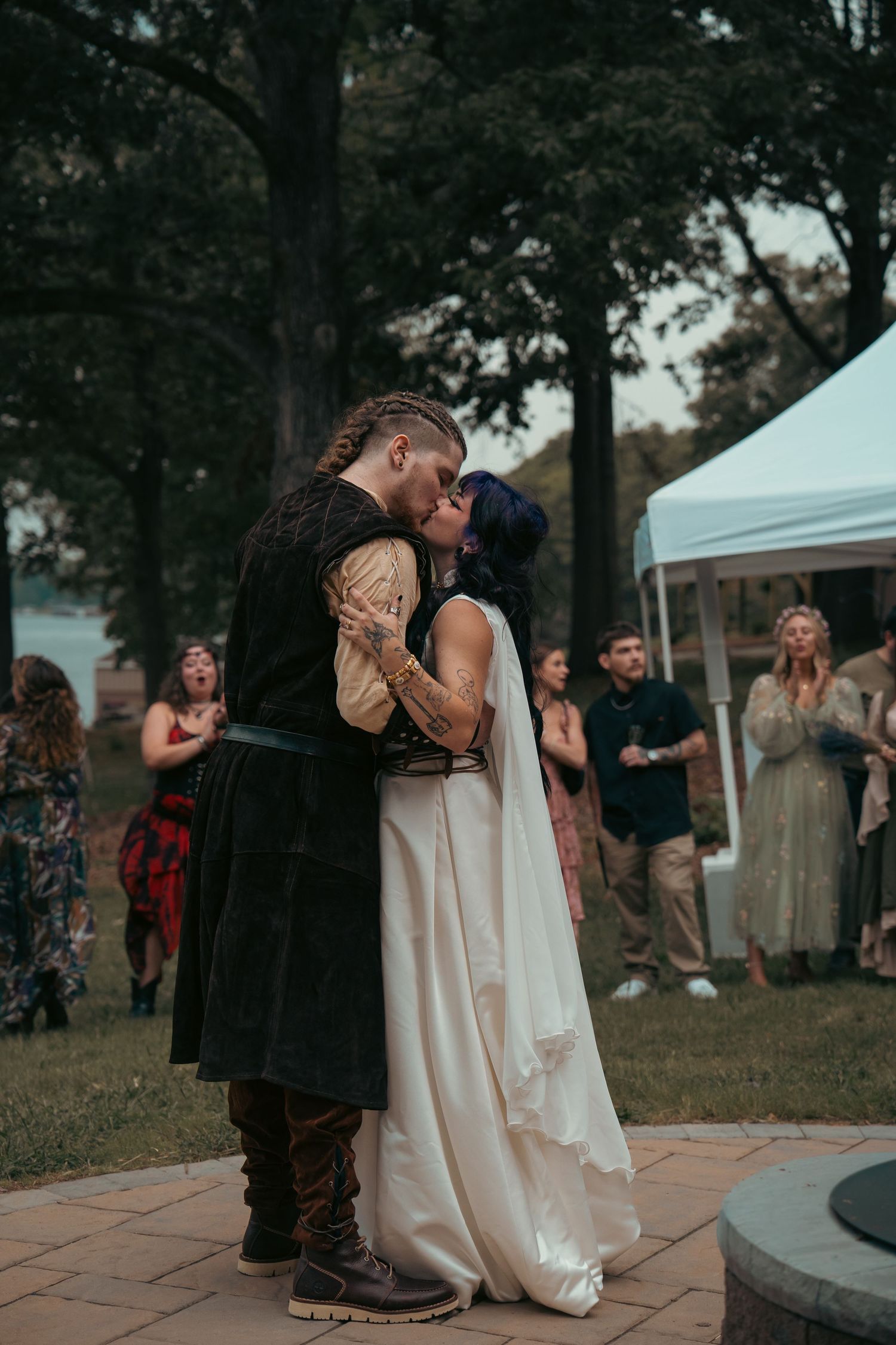 Bride and groom share first kiss in medieval wedding attire outdoors, guests applauding in background.