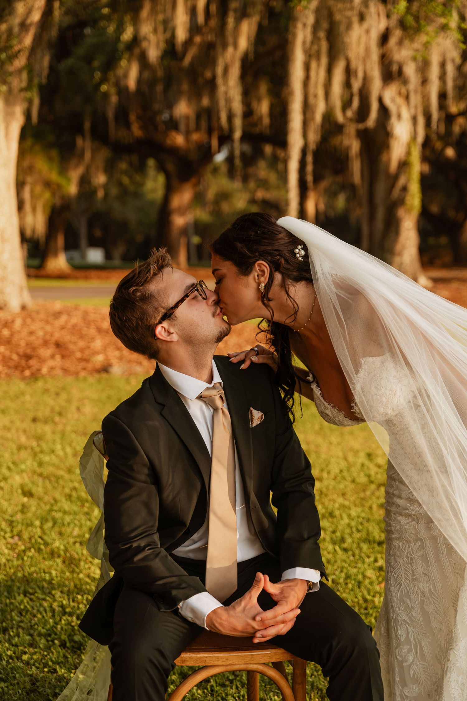Bride in white veil leans down to kiss seated groom in dark suit outdoors under Spanish moss-draped trees.
