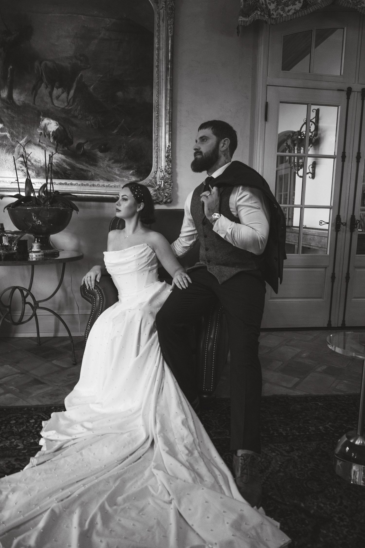 Black and white wedding photo of bride in strapless gown and bearded groom in vest posing in elegant room.