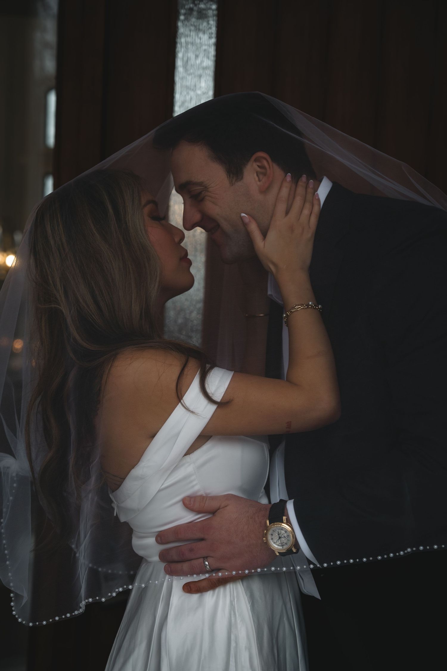 Romantic bride and groom nearly kissing under a wedding veil in a dimly lit indoor setting.