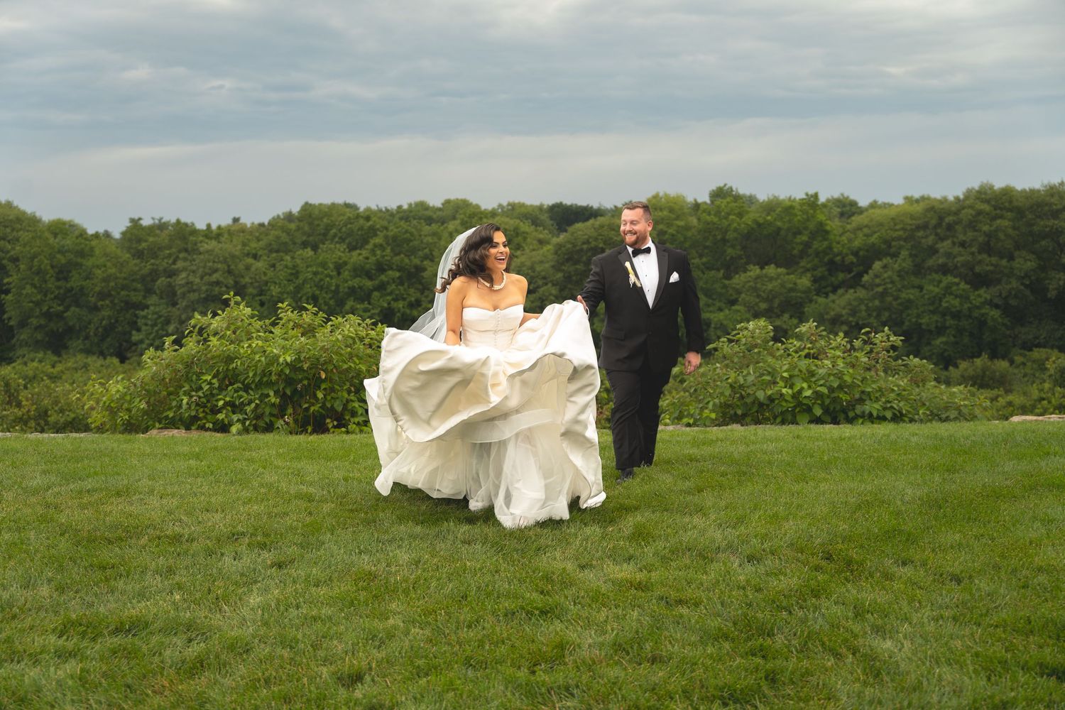 Bride in flowing white gown and groom in black tuxedo walking hand in hand on lush green lawn with trees behind.