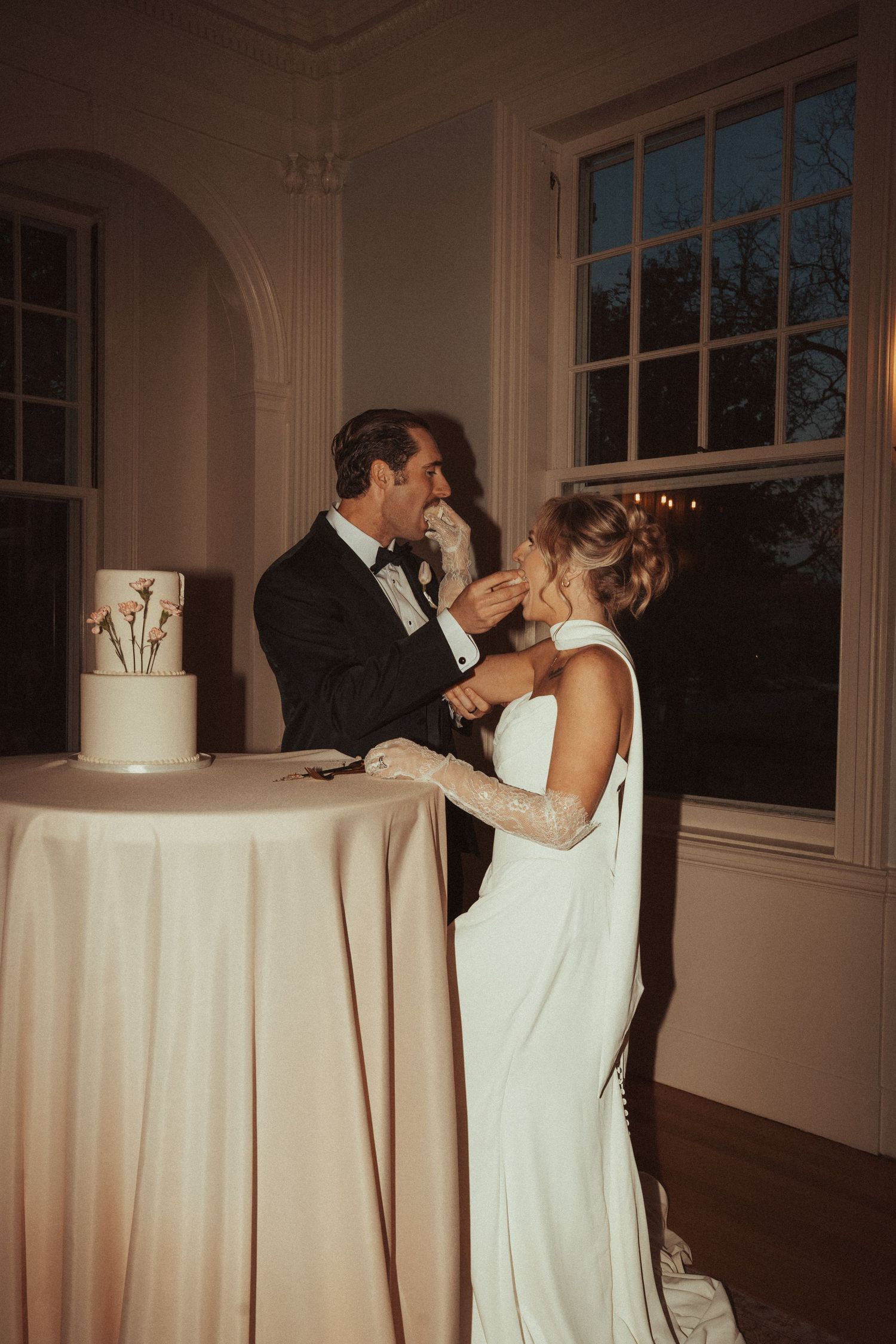 Bride and groom feeding each other wedding cake at elegant evening reception, bride in white gown with gloves.