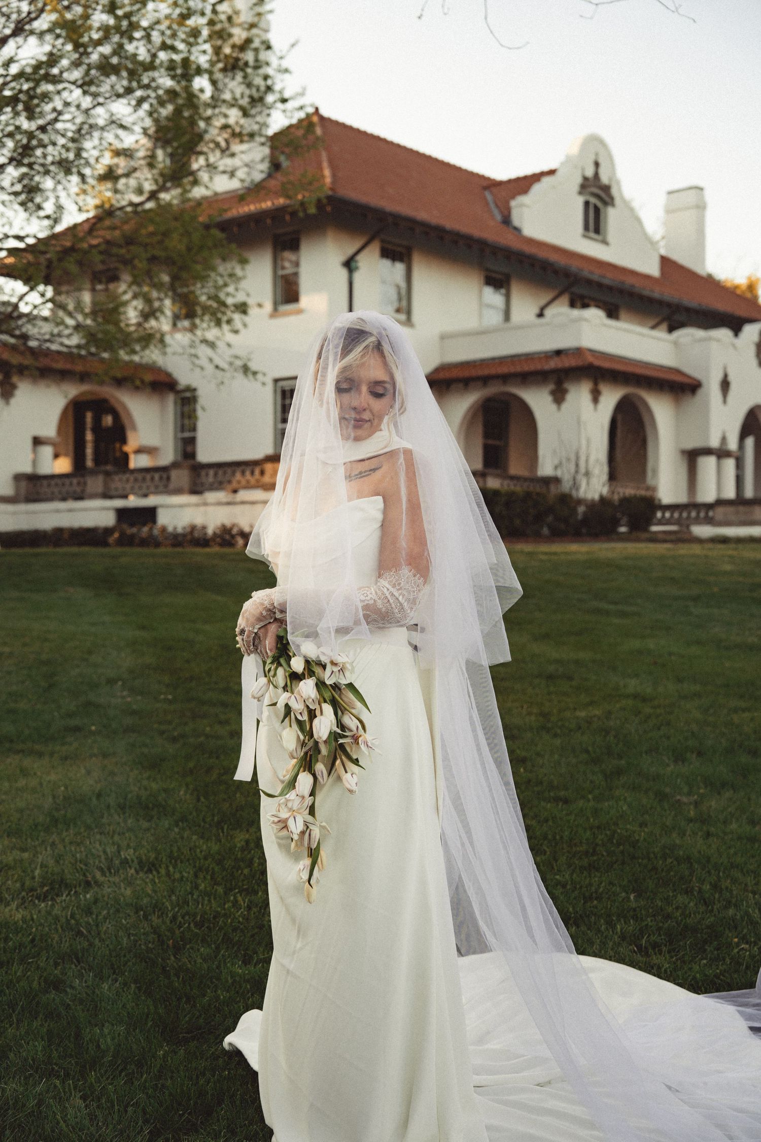 Bride in white gown and veil holds bouquet and book outside a Spanish-style mansion at dusk.