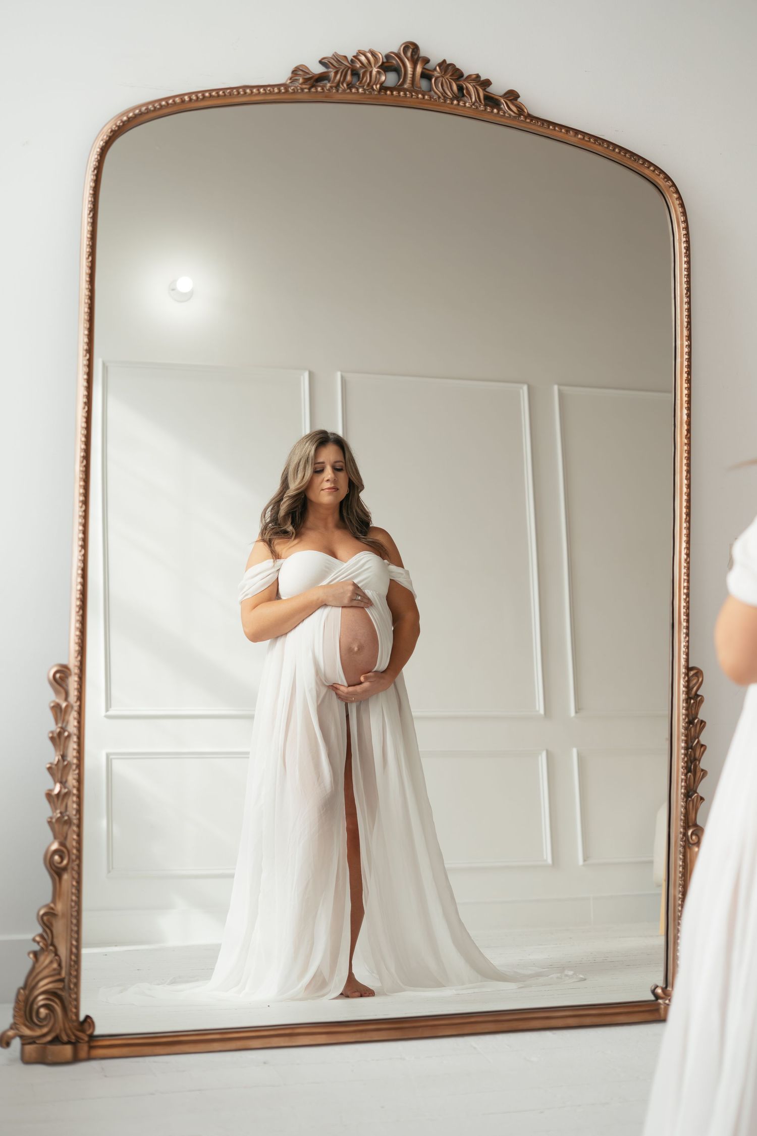 Pregnant woman in white off-shoulder gown cradling baby bump, reflected in ornate gold mirror in bright white studio.