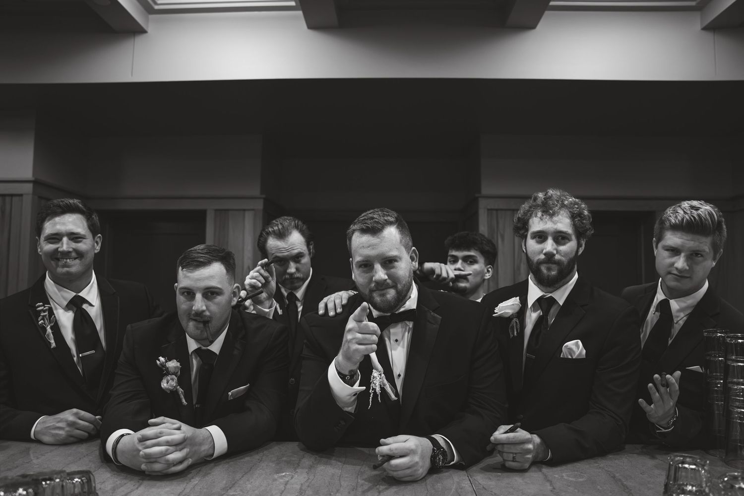 Black and white photo of a groom and groomsmen posing at a bar, raising glasses in a celebratory toast.