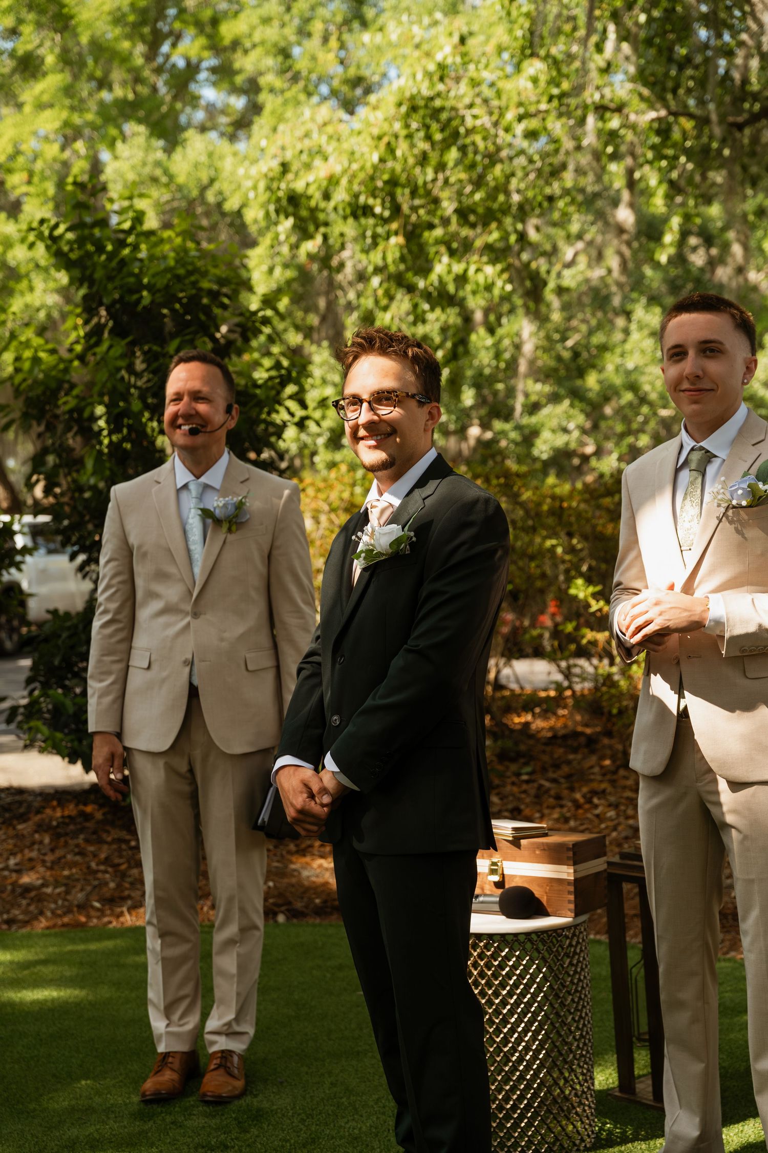 Groom in black suit smiles at altar flanked by groomsmen in tan suits during outdoor wedding ceremony.