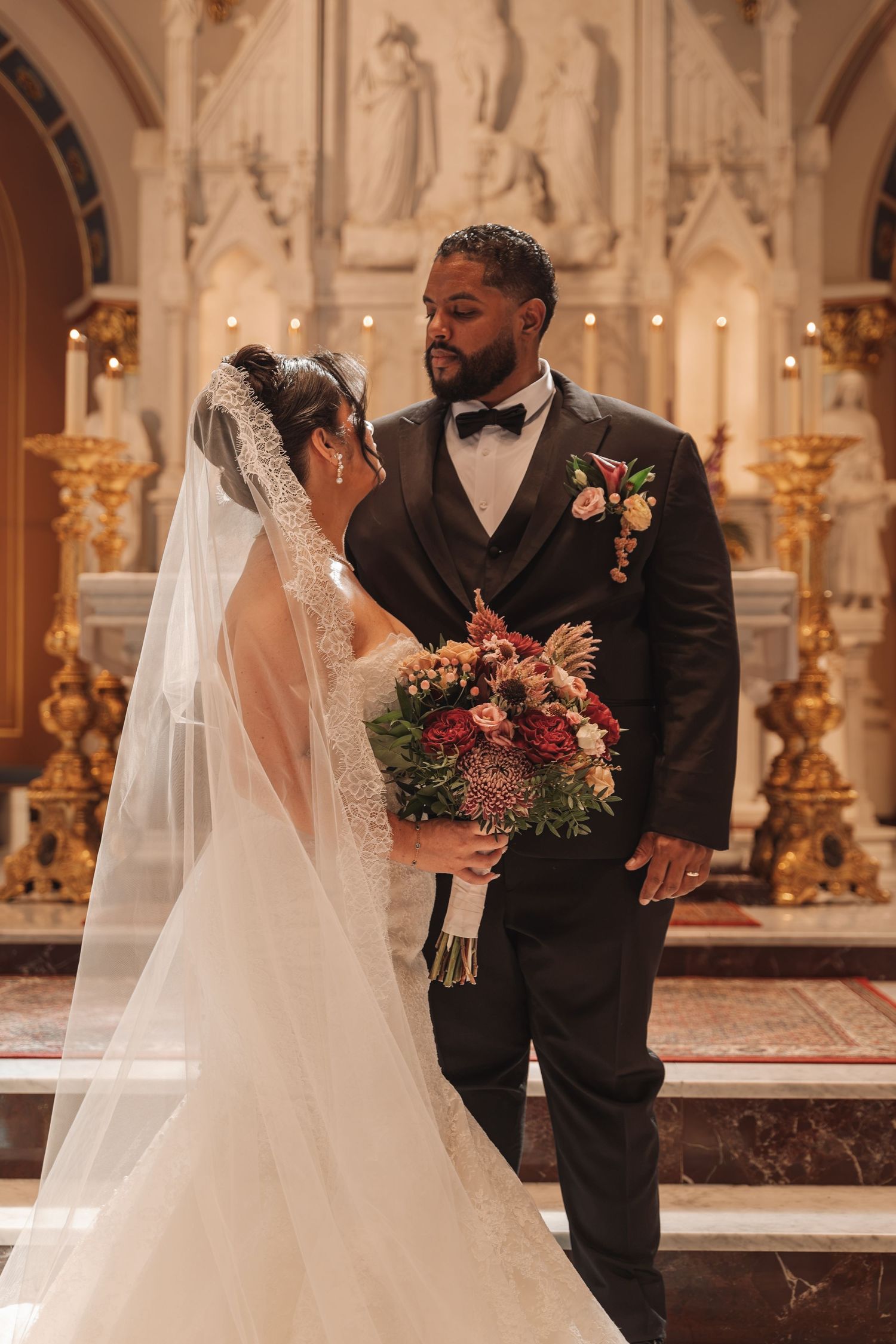 Bride and groom share an intimate moment at church altar, bride holding red and pink bouquet in elegant lace gown and veil.