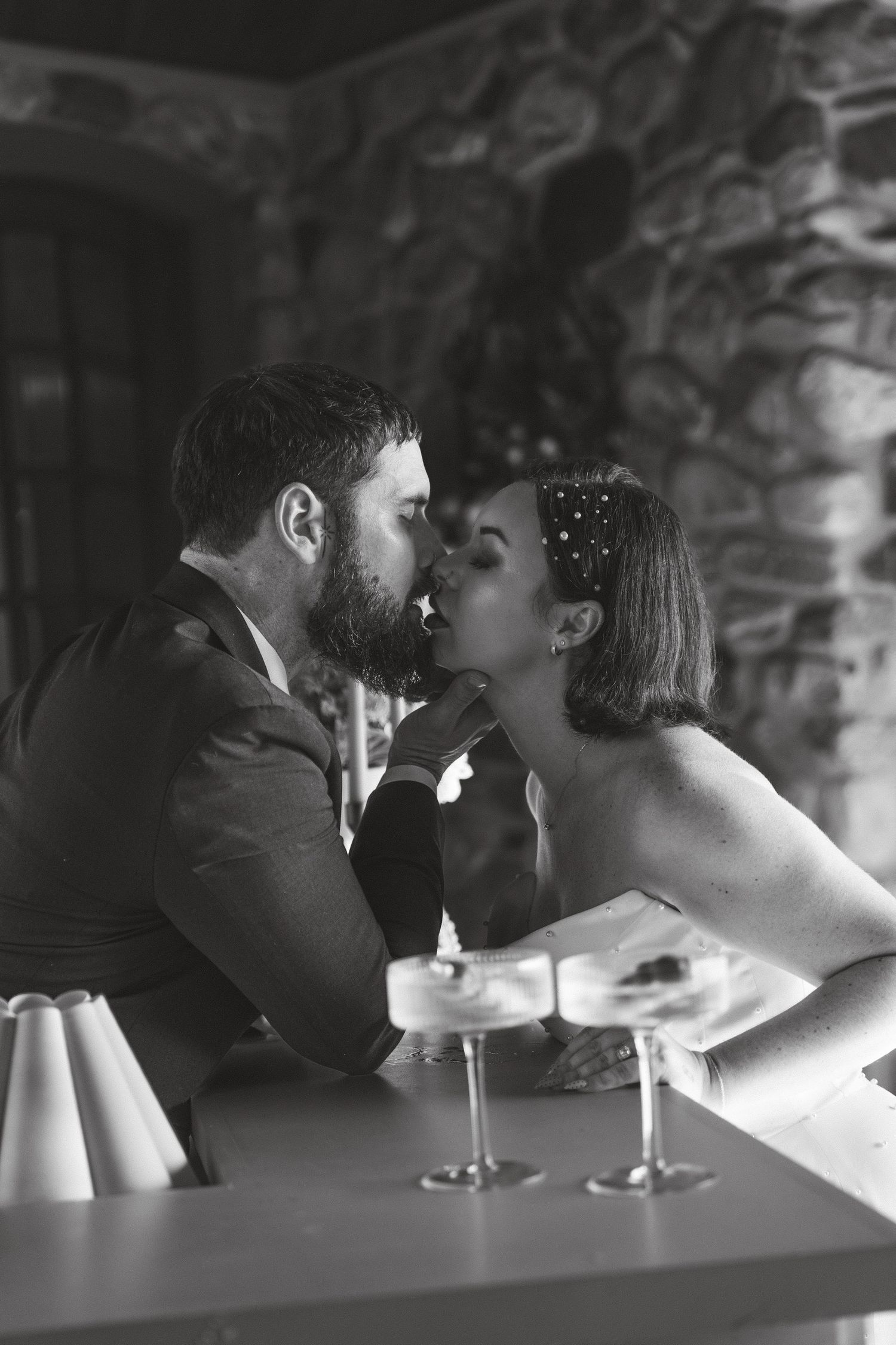 Black and white photo of bride and groom sharing a romantic kiss at a bar with champagne glasses.