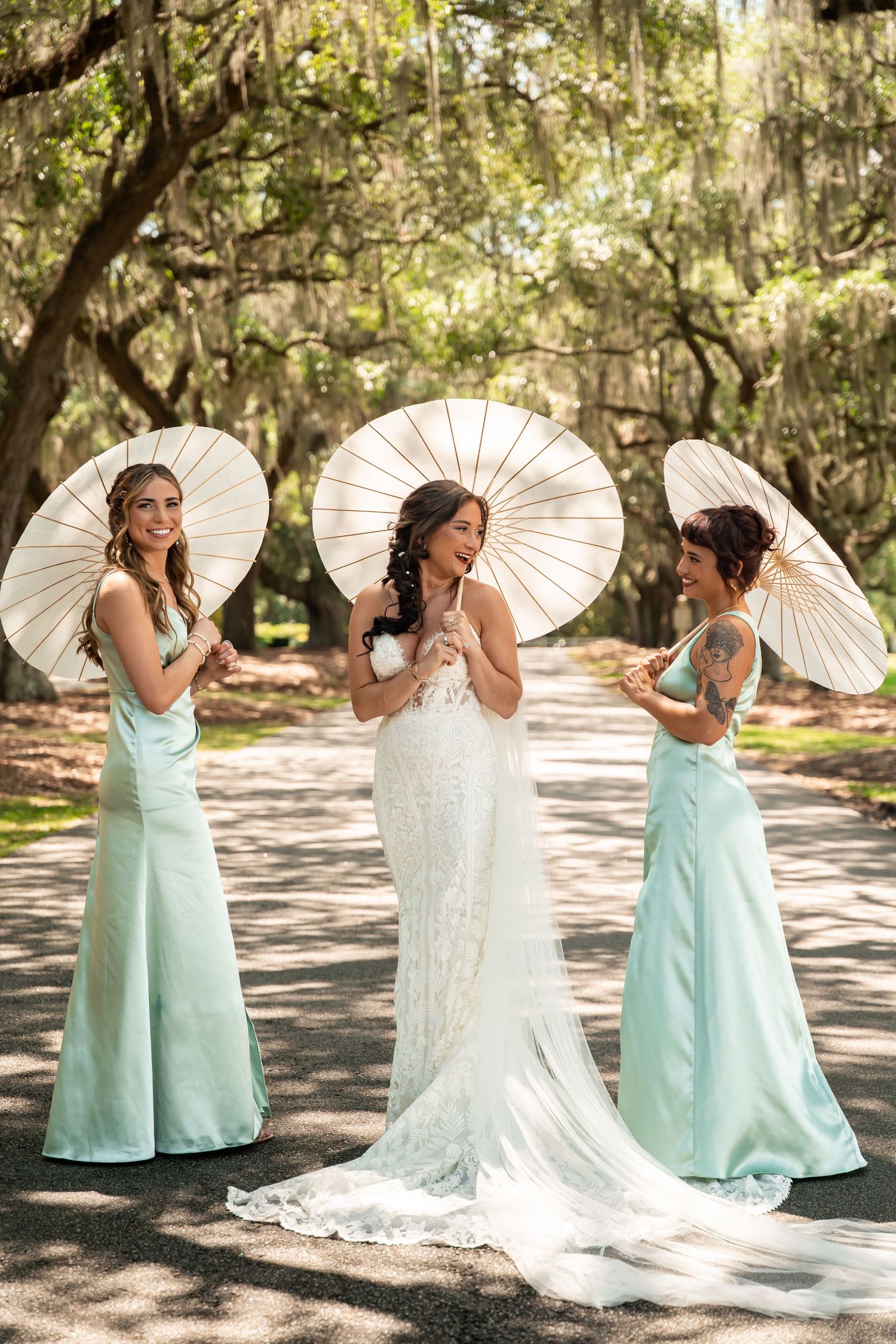 Bride in white lace gown with two bridesmaids in mint dresses holding white parasols on a tree-lined path.