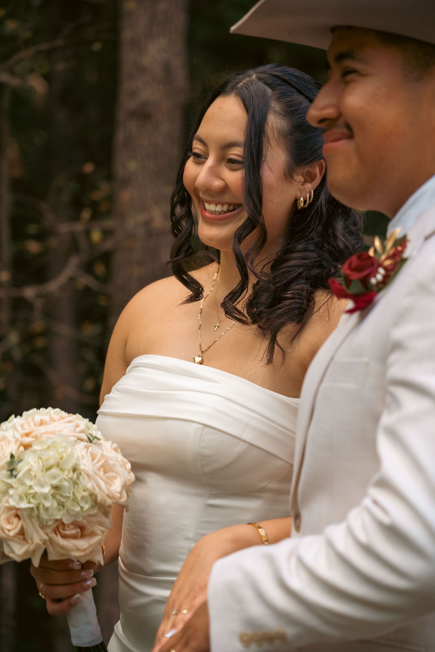 Smiling bride in white strapless gown holds cream bouquet beside groom in white suit and cowboy hat outdoors.
