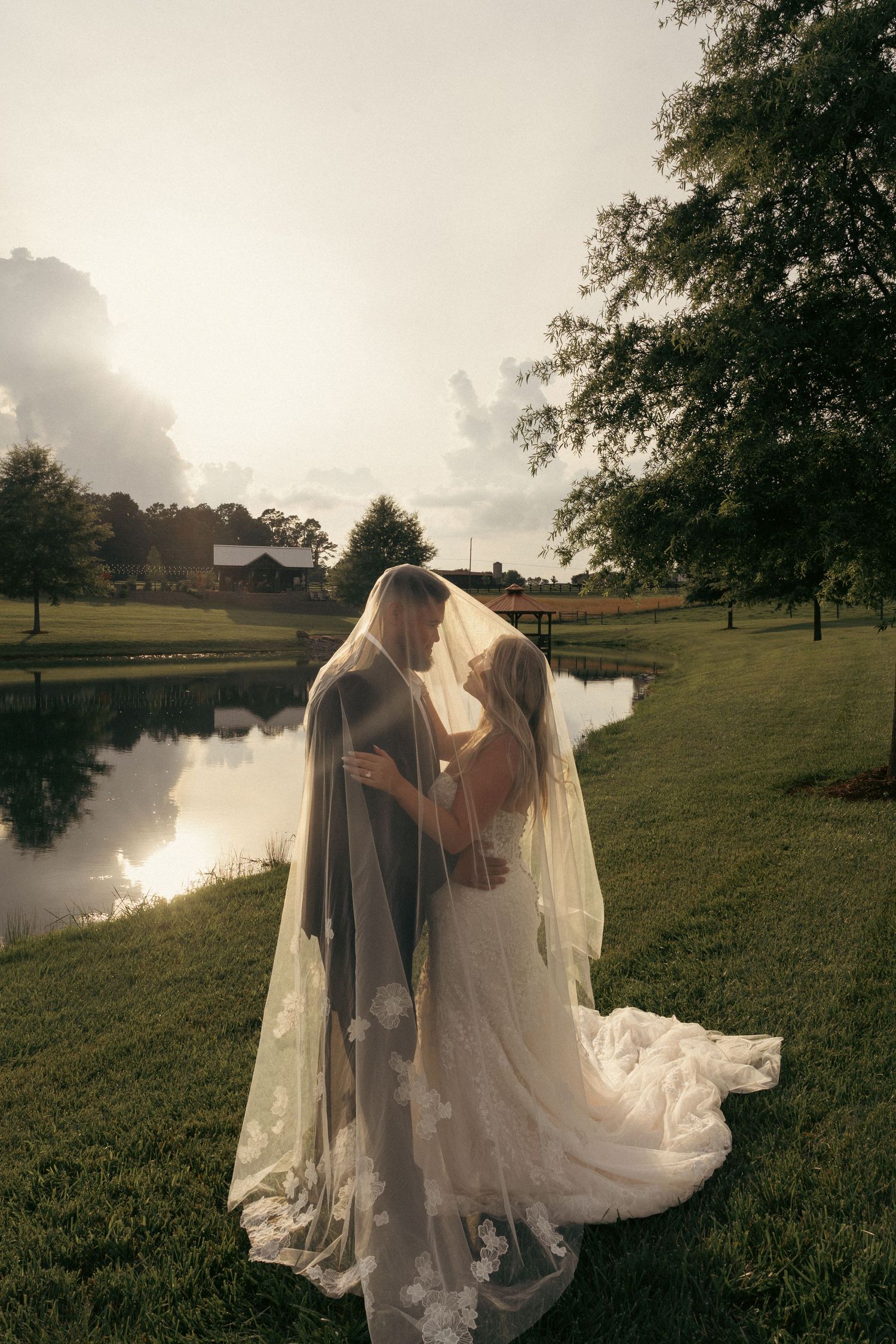 Bride and groom kiss under flowing lace veil beside a serene pond at golden hour during outdoor wedding.