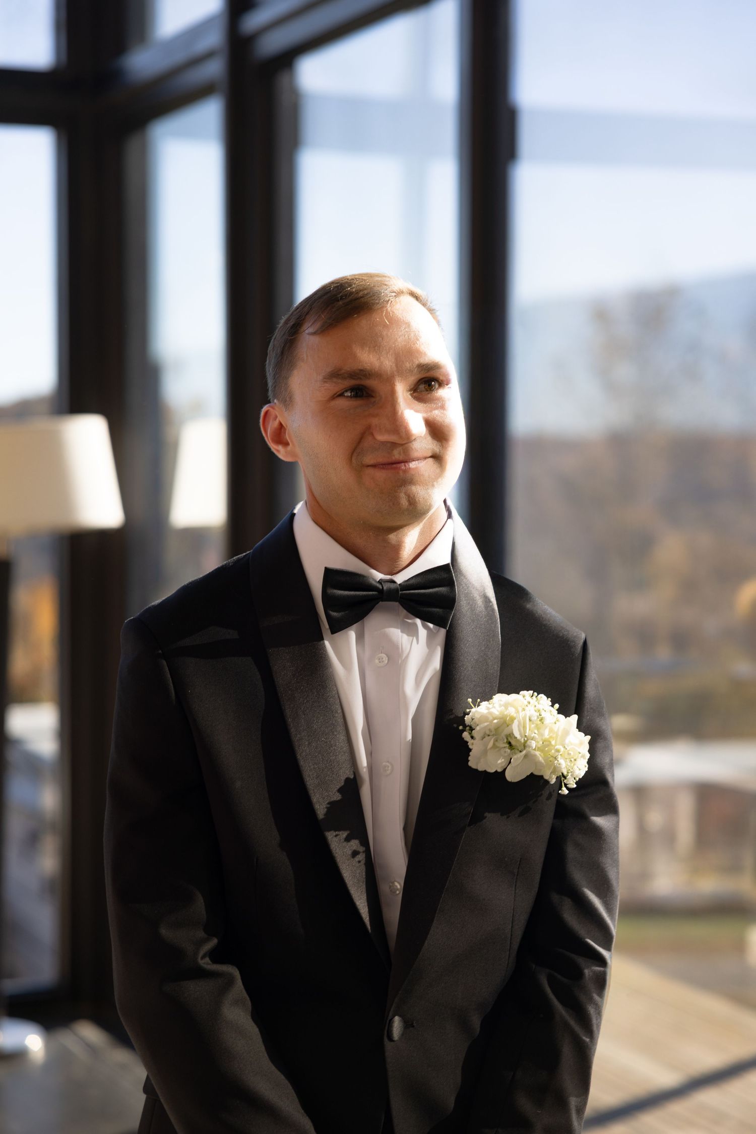 Smiling groom in black tuxedo with bow tie and white boutonniere stands near large windows on wedding day.