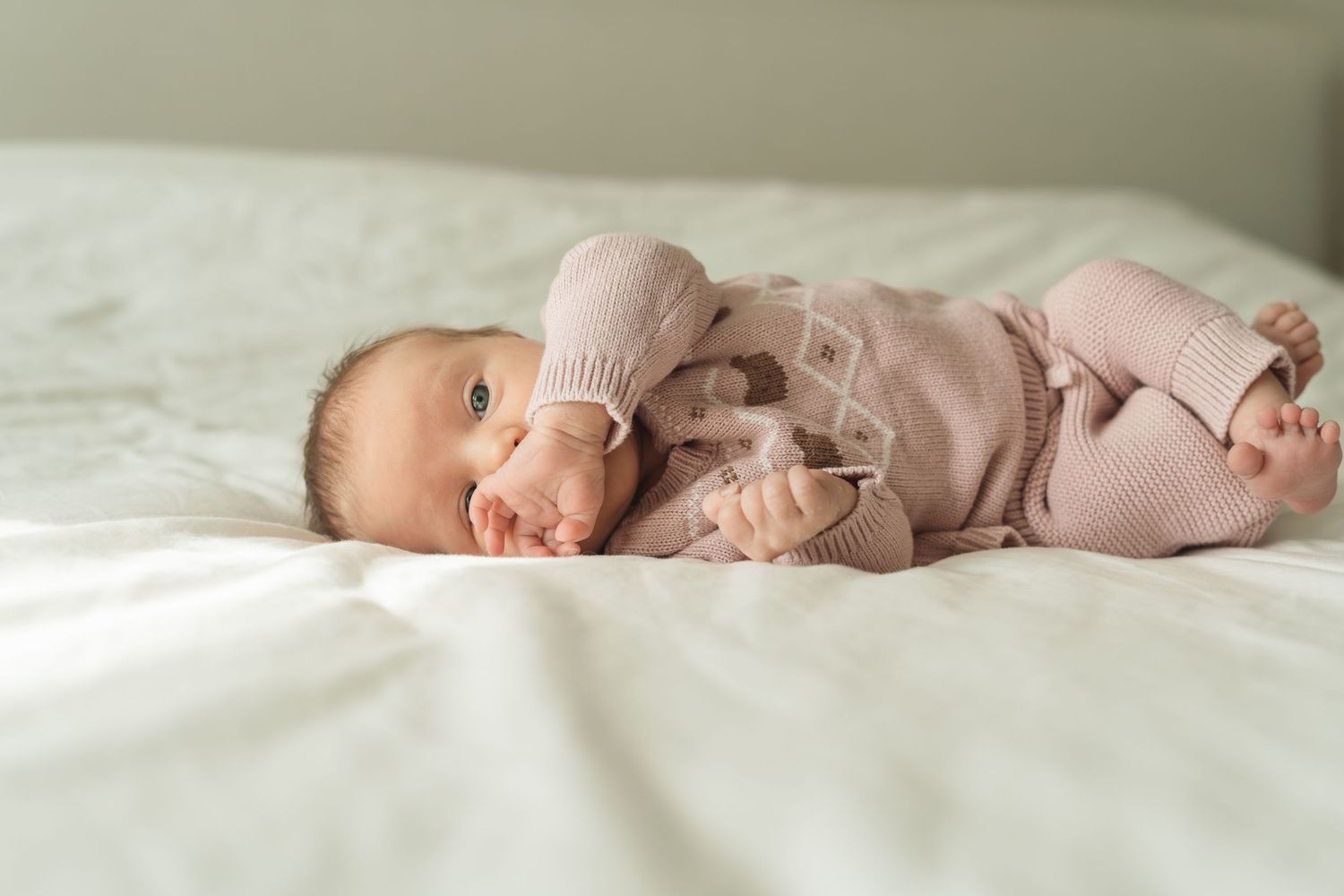 Adorable baby in pink knit outfit lying on white bedding, playfully holding feet up in the air.