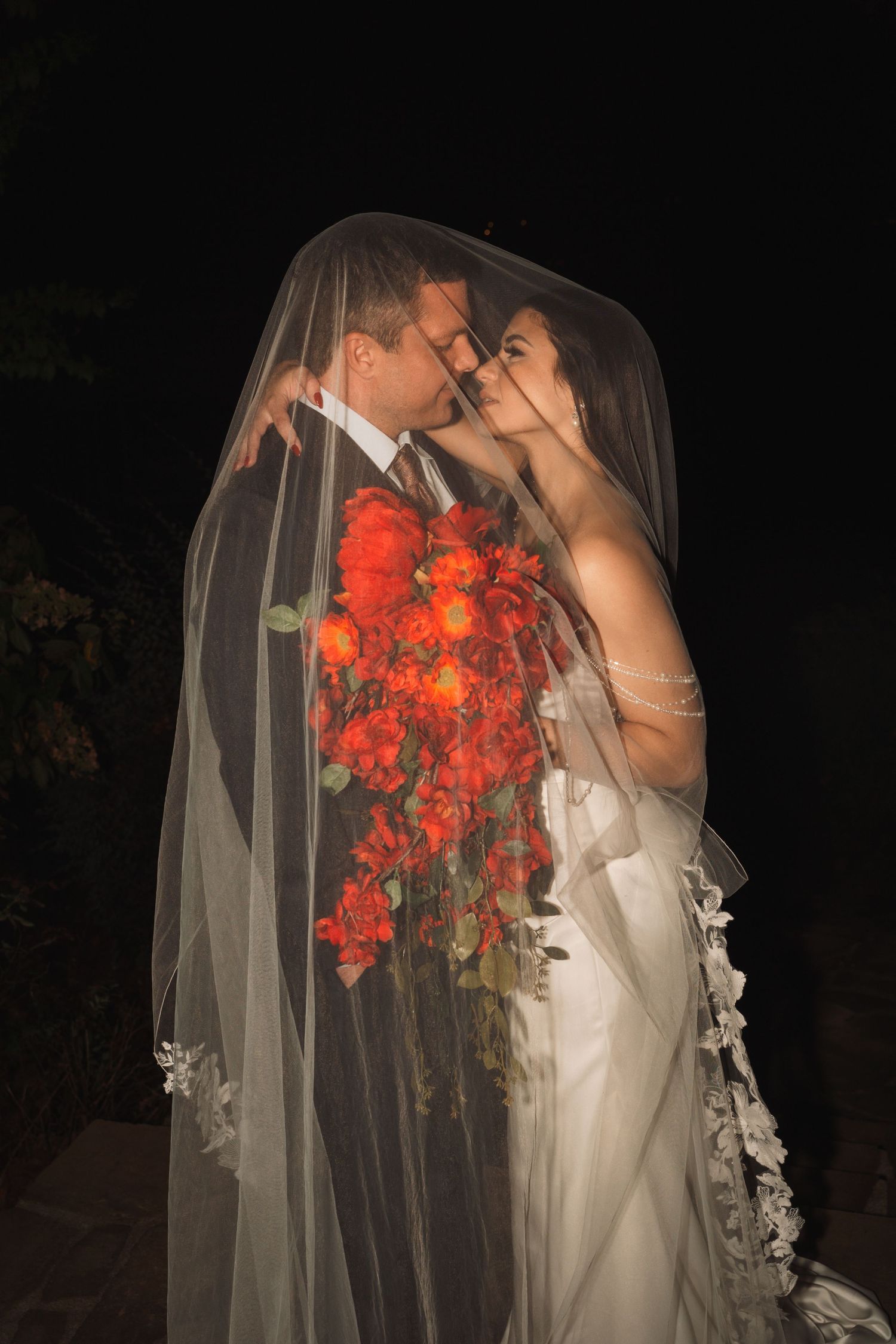 Bride and groom share an intimate moment under a sheer lace-trimmed veil at night, holding a cascading bouquet of red-orange roses.