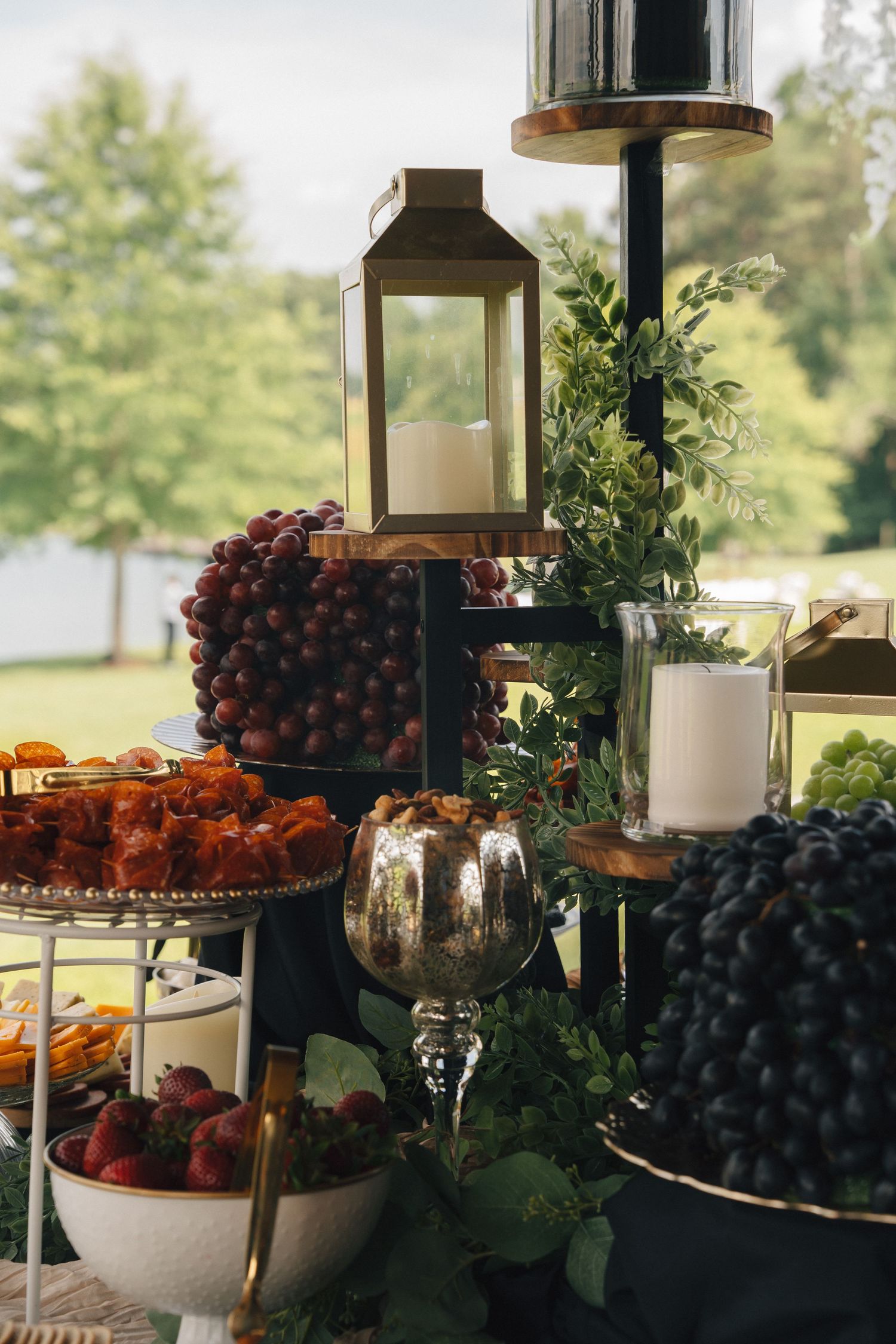 An elegant wedding charcuterie display with grapes, strawberries, and a gold lantern centerpiece outdoors.