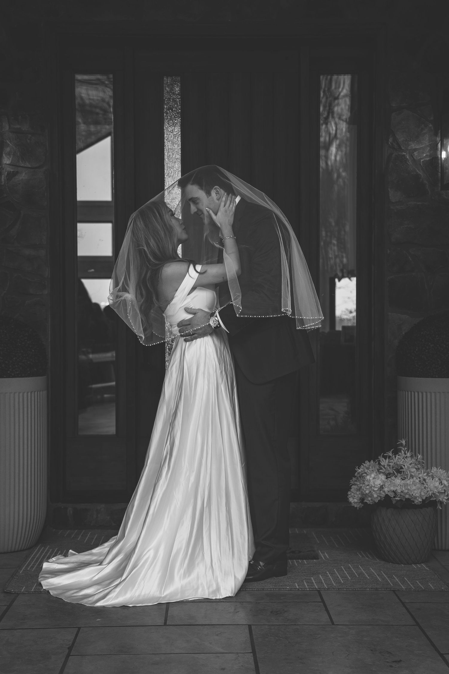 Black and white photo of bride and groom sharing a kiss under a veil at a dark wooden door.