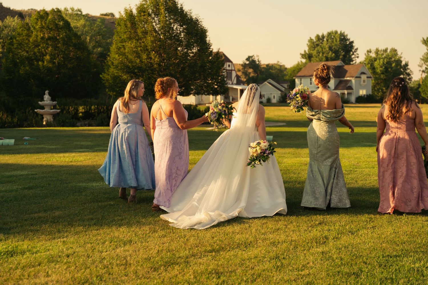 Bride in white gown stands with bridesmaids in colorful dresses on green lawn at golden hour wedding celebration.