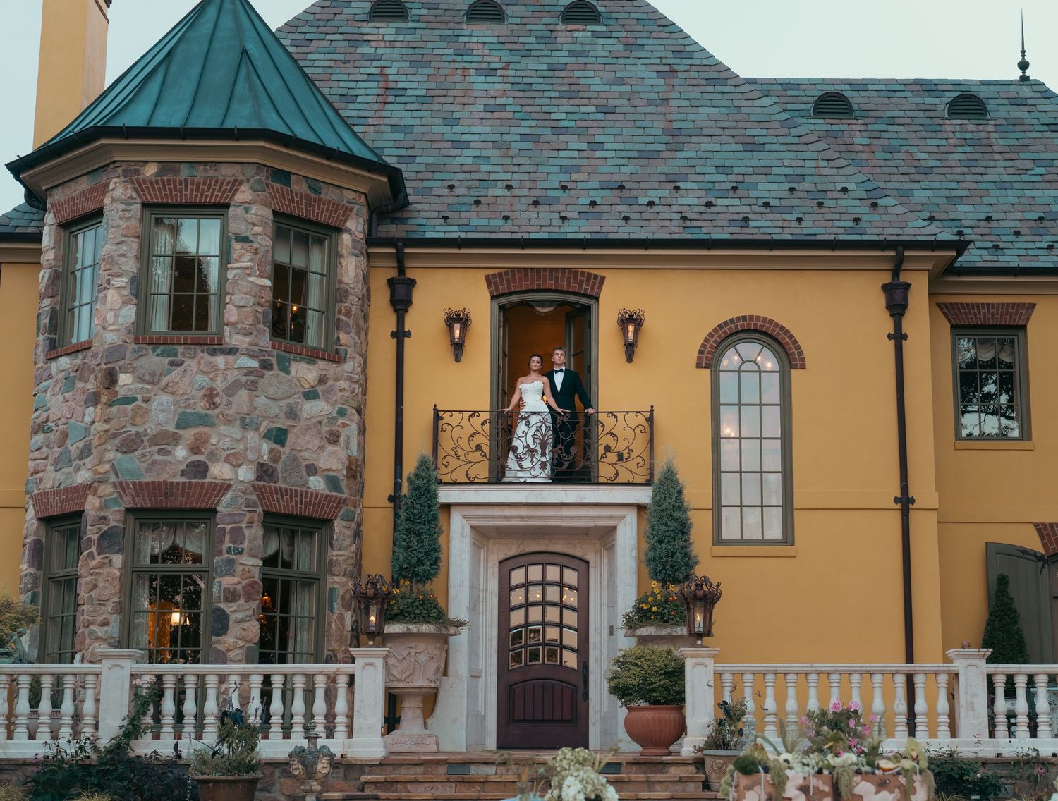 Couple poses on balcony of elegant stone and yellow stucco mansion with turret and wrought iron railing.
