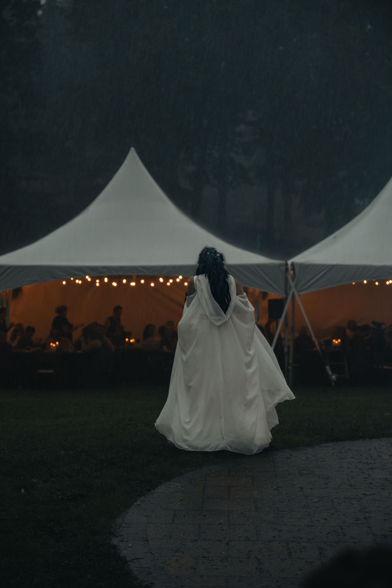 Bride in white gown and cape walks toward glowing wedding tent with string lights at dusk.